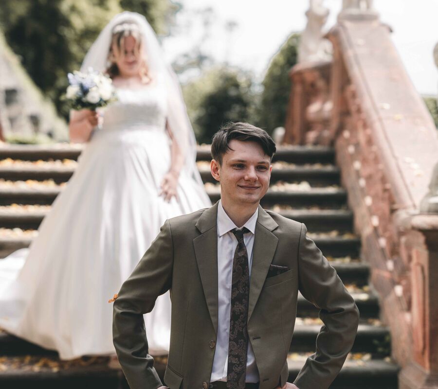 First Look auf einer barocken Steintreppe: Ein lächelnder Bräutigam im olivgrünen Hochzeitsanzug wartet gespannt, während seine Braut im weißen Hochzeitskleid mit Schleier hinter ihm die Stufen herabsteigt.