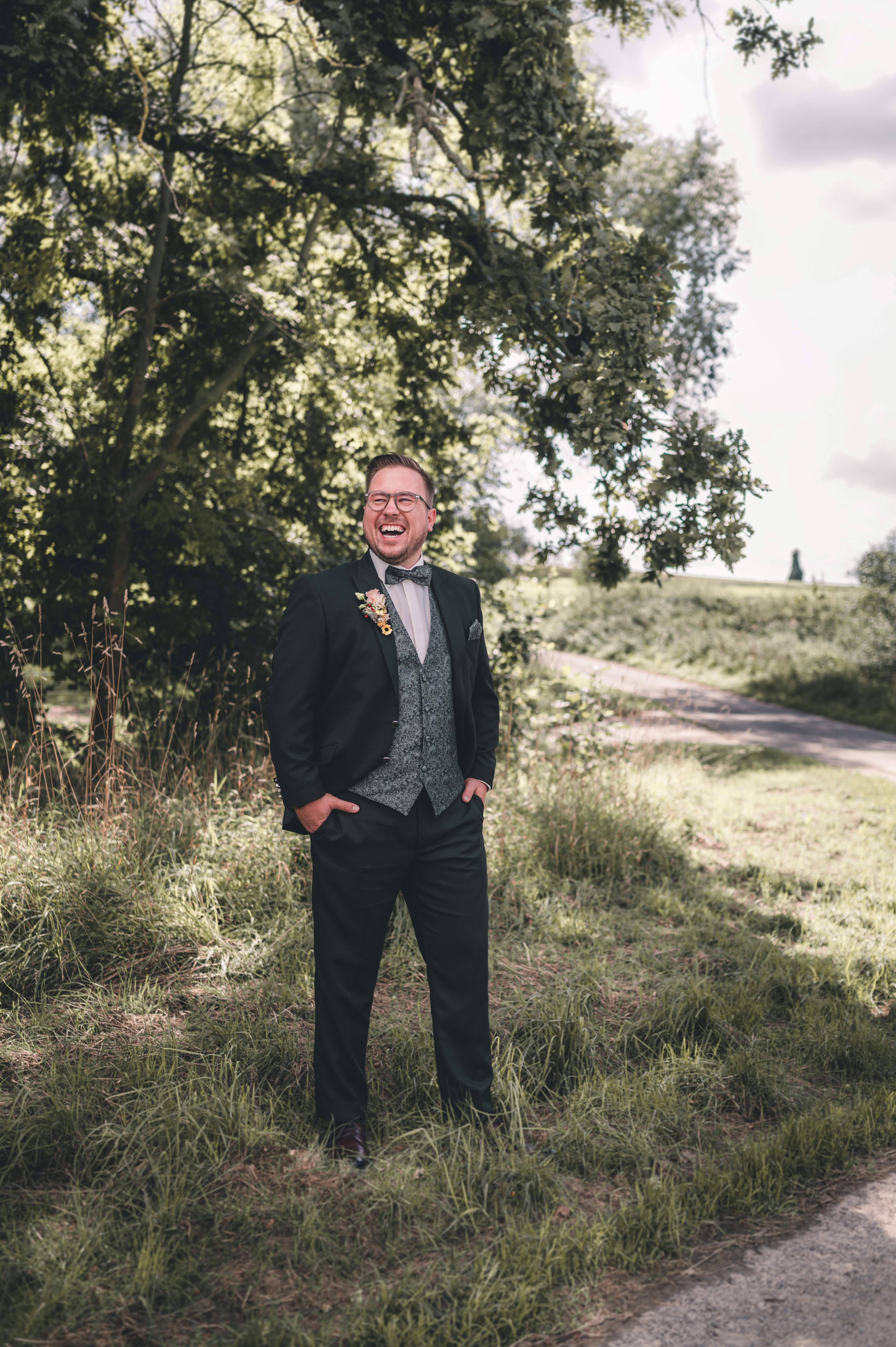 Der Bräutigam im gemusterten, dunkelgrünen Hochzeitsanzug steht mit den Händen in den Taschen im Wald und lacht laut und unbeschwert.