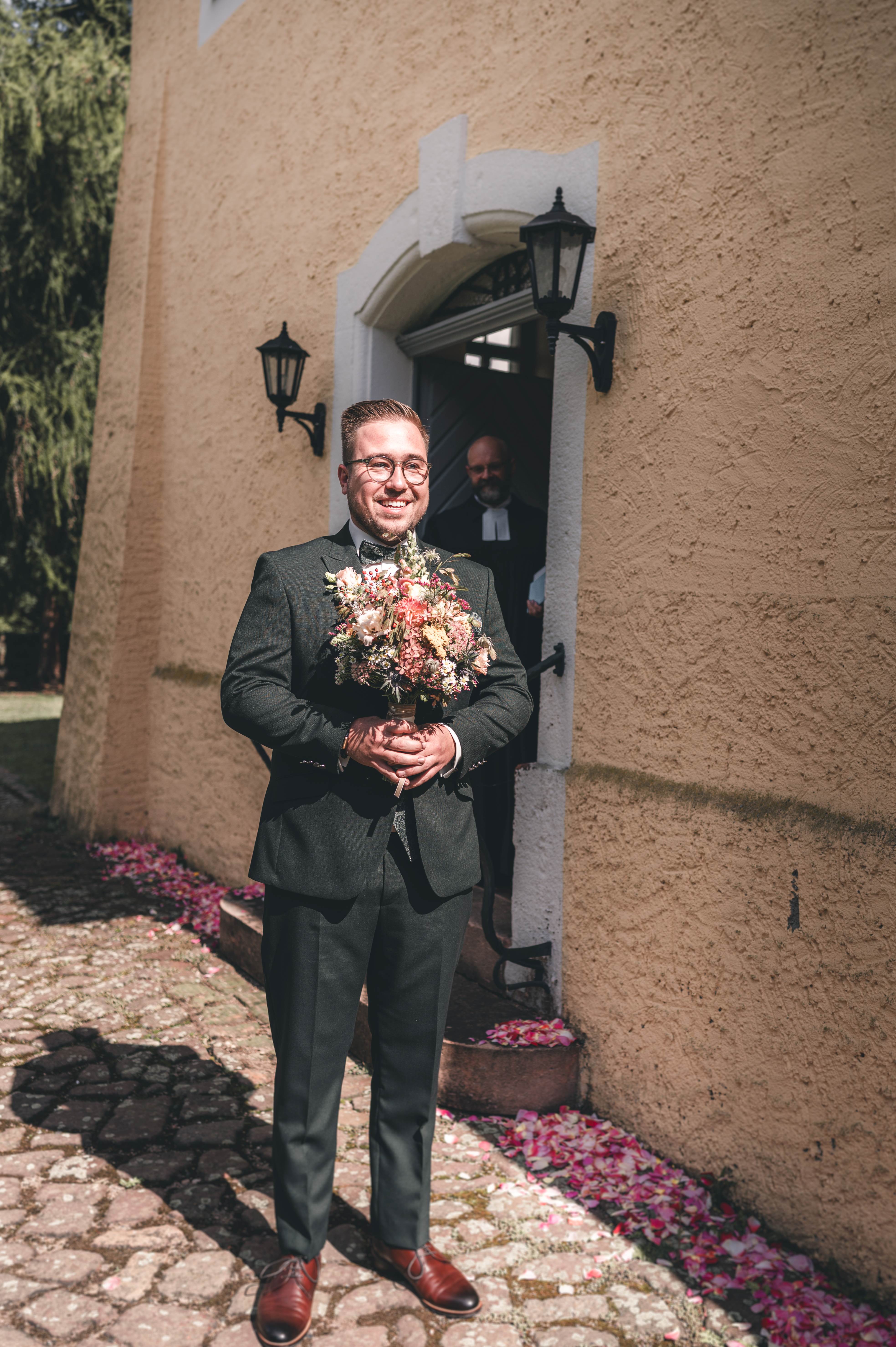 Der Bräutigam steht im eleganten dunkelgrünen Anzug lächelnd mit dem bunten Wiesenblumenstrauß vor dem Portal der Kirche in Leisnig.