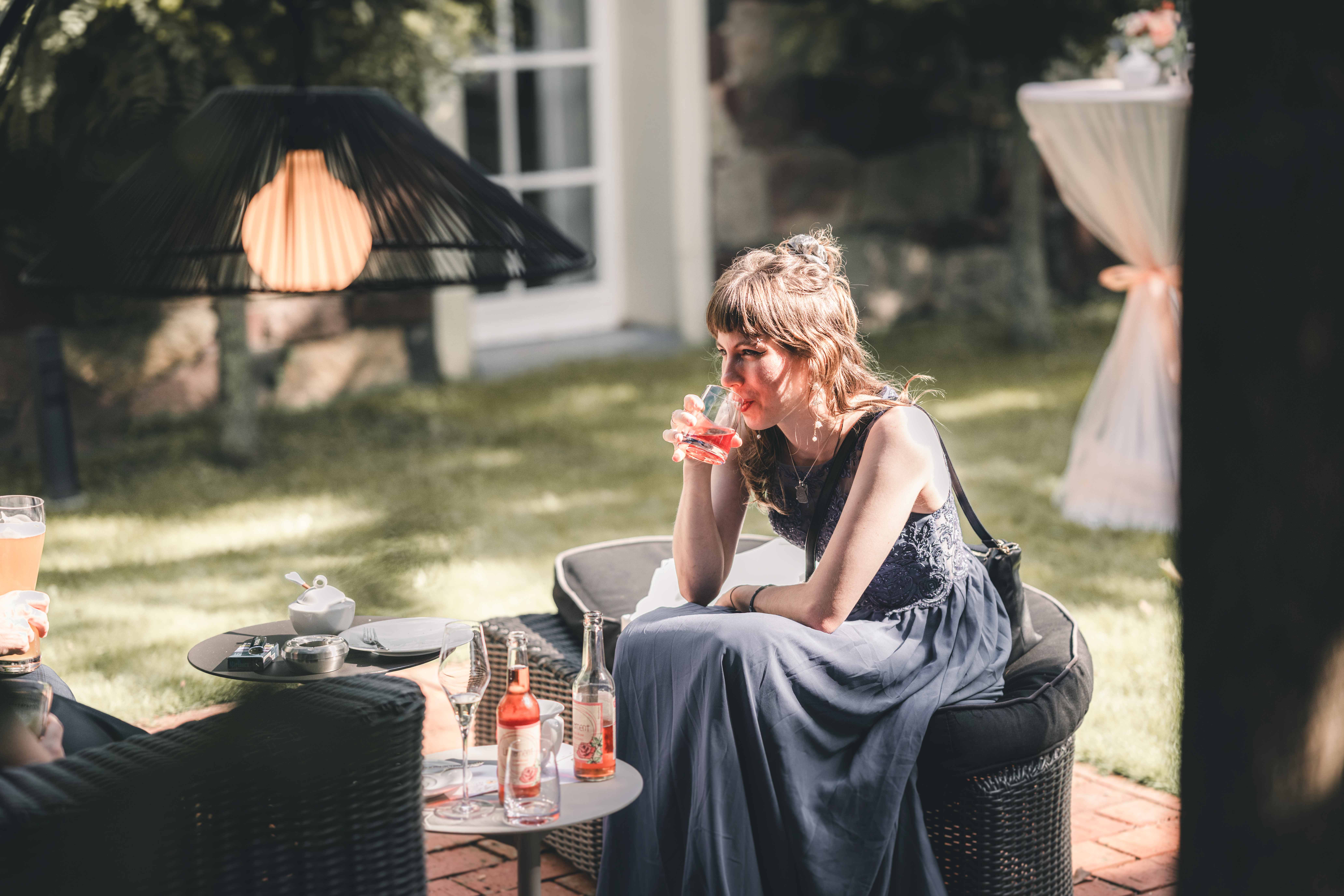 Eine Hochzeitsgästin im eleganten blauen Kleid sitzt entspannt in einer Rattan-Lounge im Garten und genießt einen kühlen Drink.