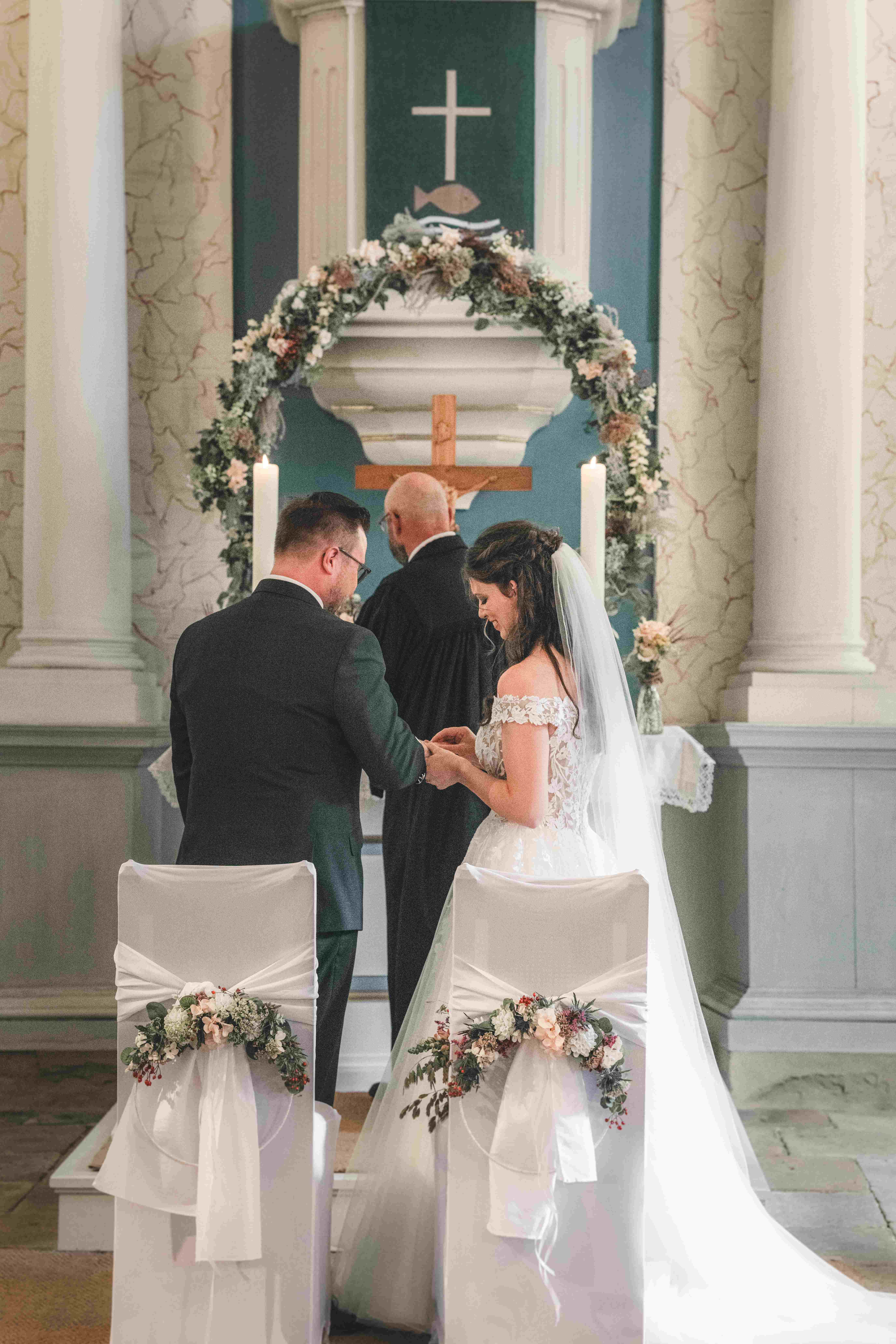 Der Bräutigam steckt seiner Braut vor dem mit einem Blumenbogen verzierten Altar den Ehering an den Finger.