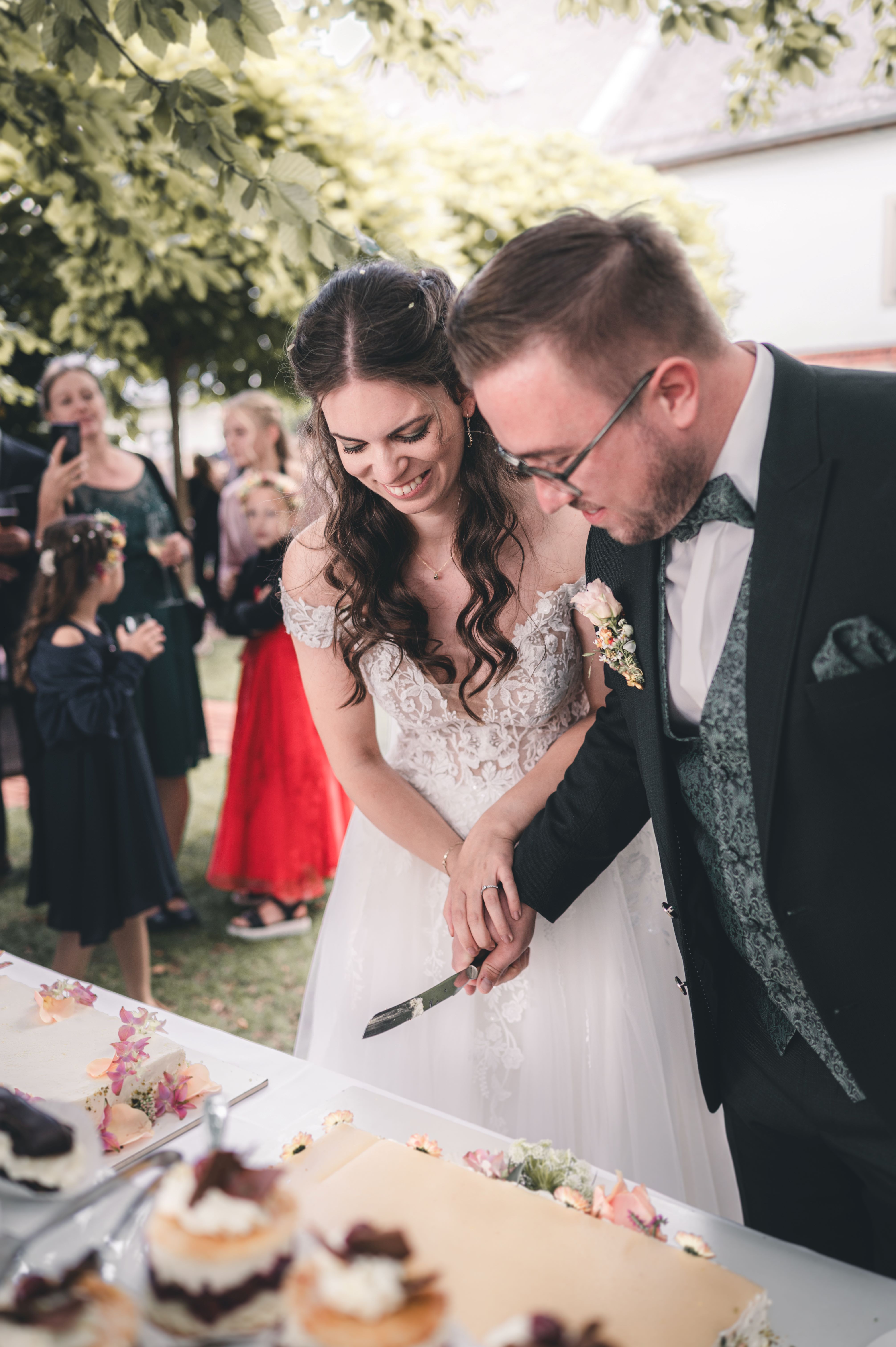 Das Brautpaar hält gemeinsam das Messer und schneidet lächelnd die festliche Hochzeitstorte unter den Bäumen am Hofgut Munz an.