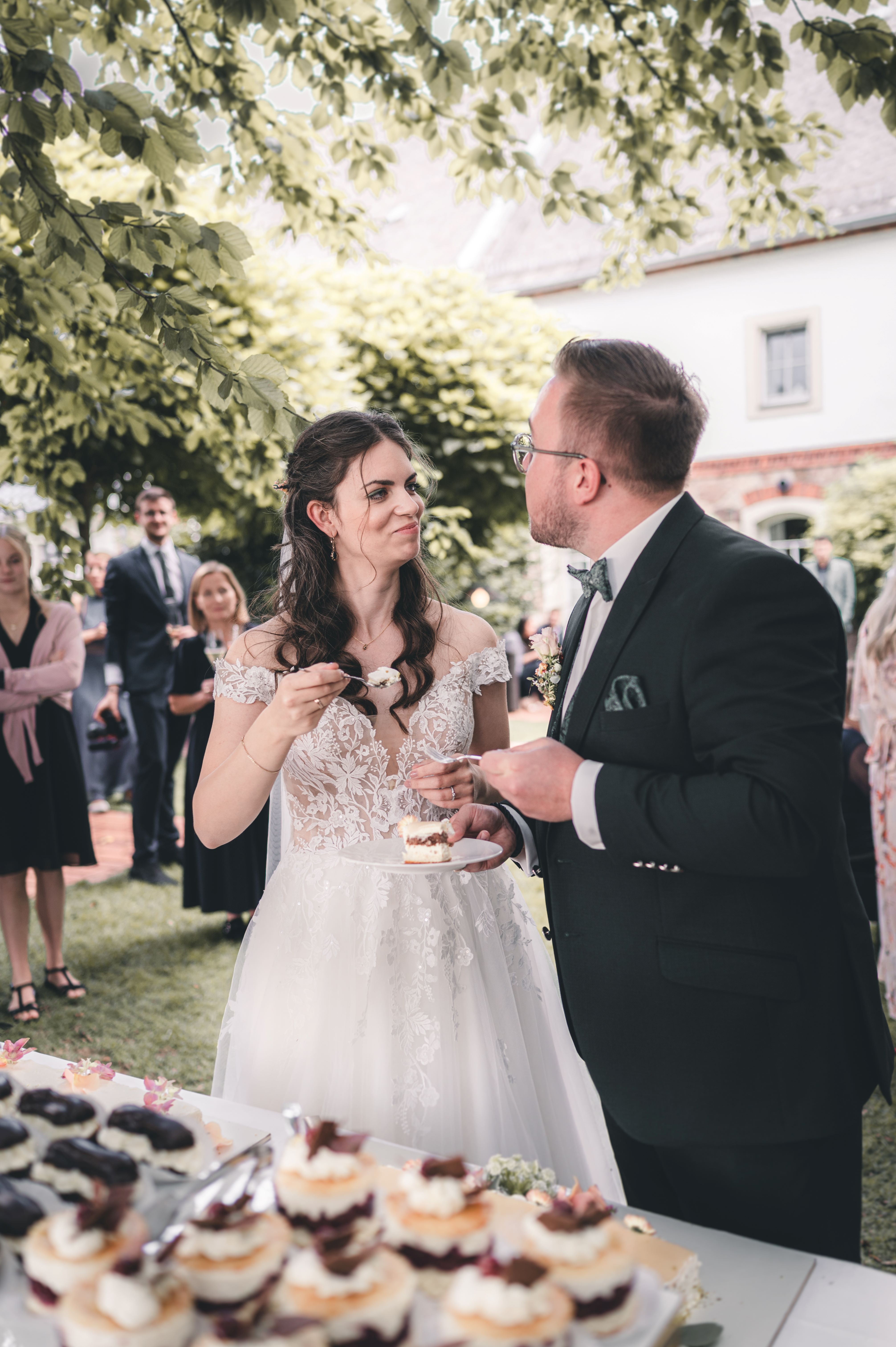 Das Brautpaar füttert sich am Nachmittag im Garten des Hofguts Munz in Leisnig lachend gegenseitig mit einem Stück Hochzeitstorte.