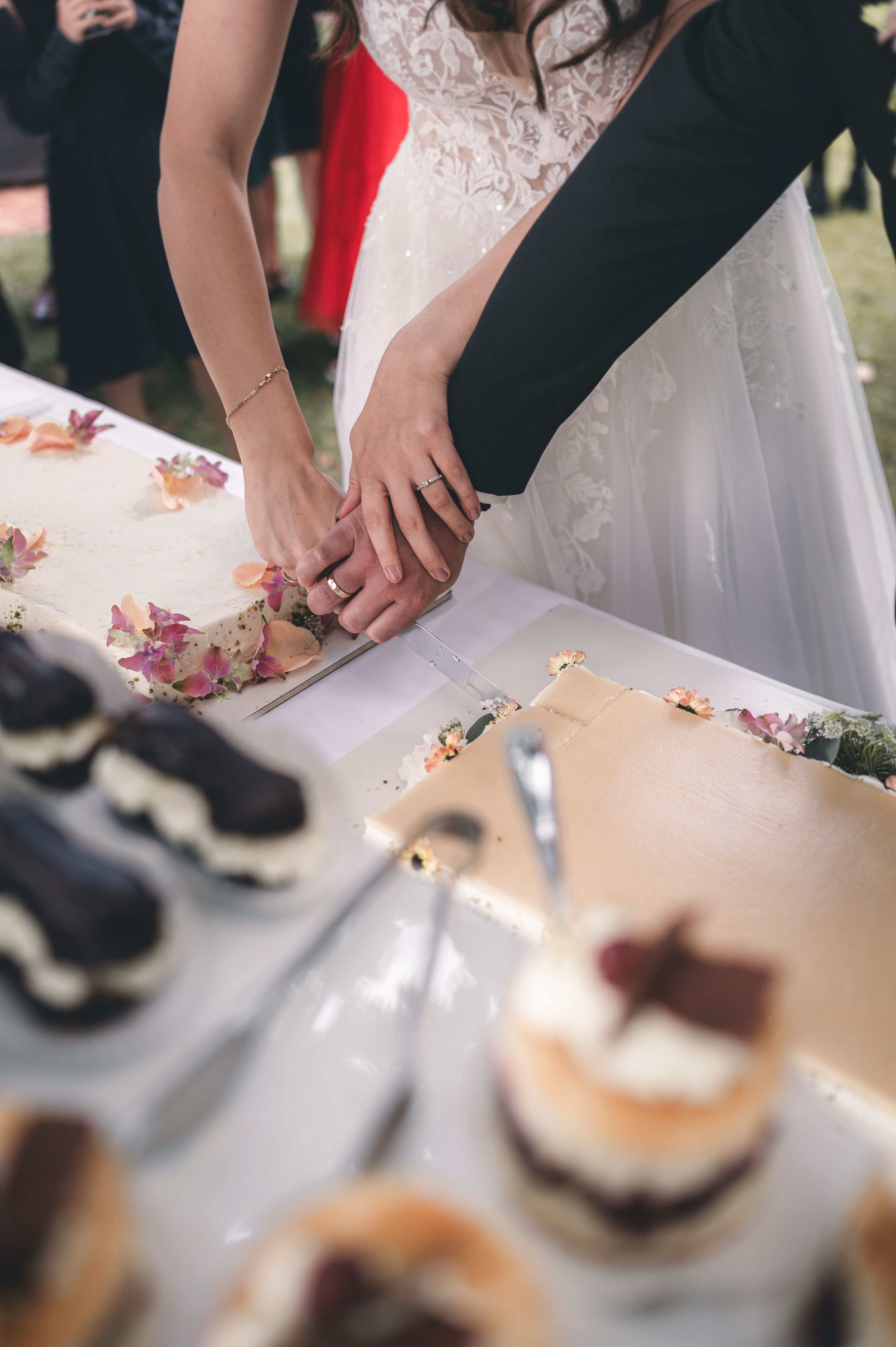 Nahaufnahme der Hände des Brautpaares mit ihren funkelnden Eheringen, wie sie gemeinsam das Messer durch die Torte führen.