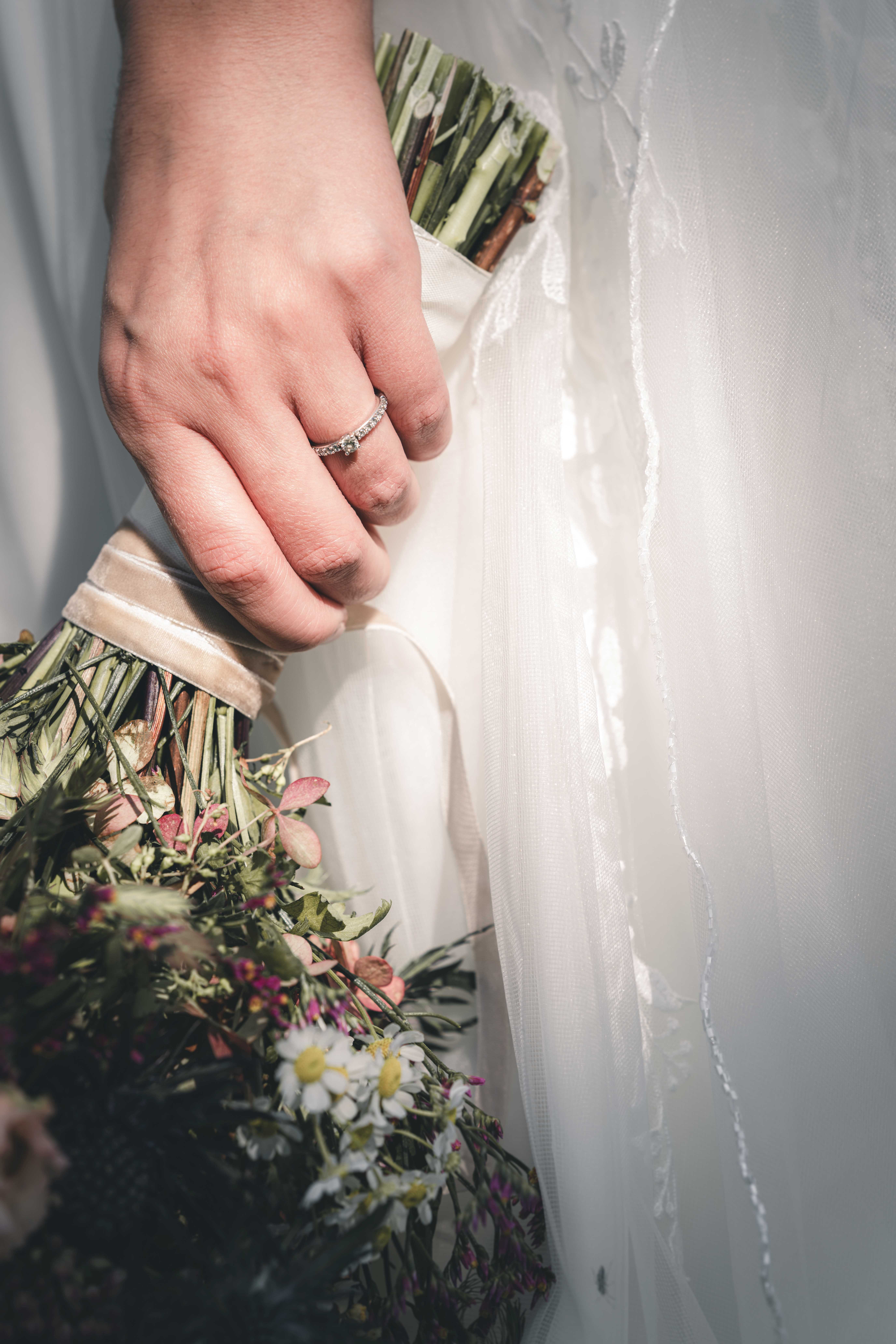 Nahaufnahme der Hand der Braut mit einem funkelnden Diamantring, die fest den mit Samtband gewickelten Stiel ihres Wiesenblumenstraußes hält.