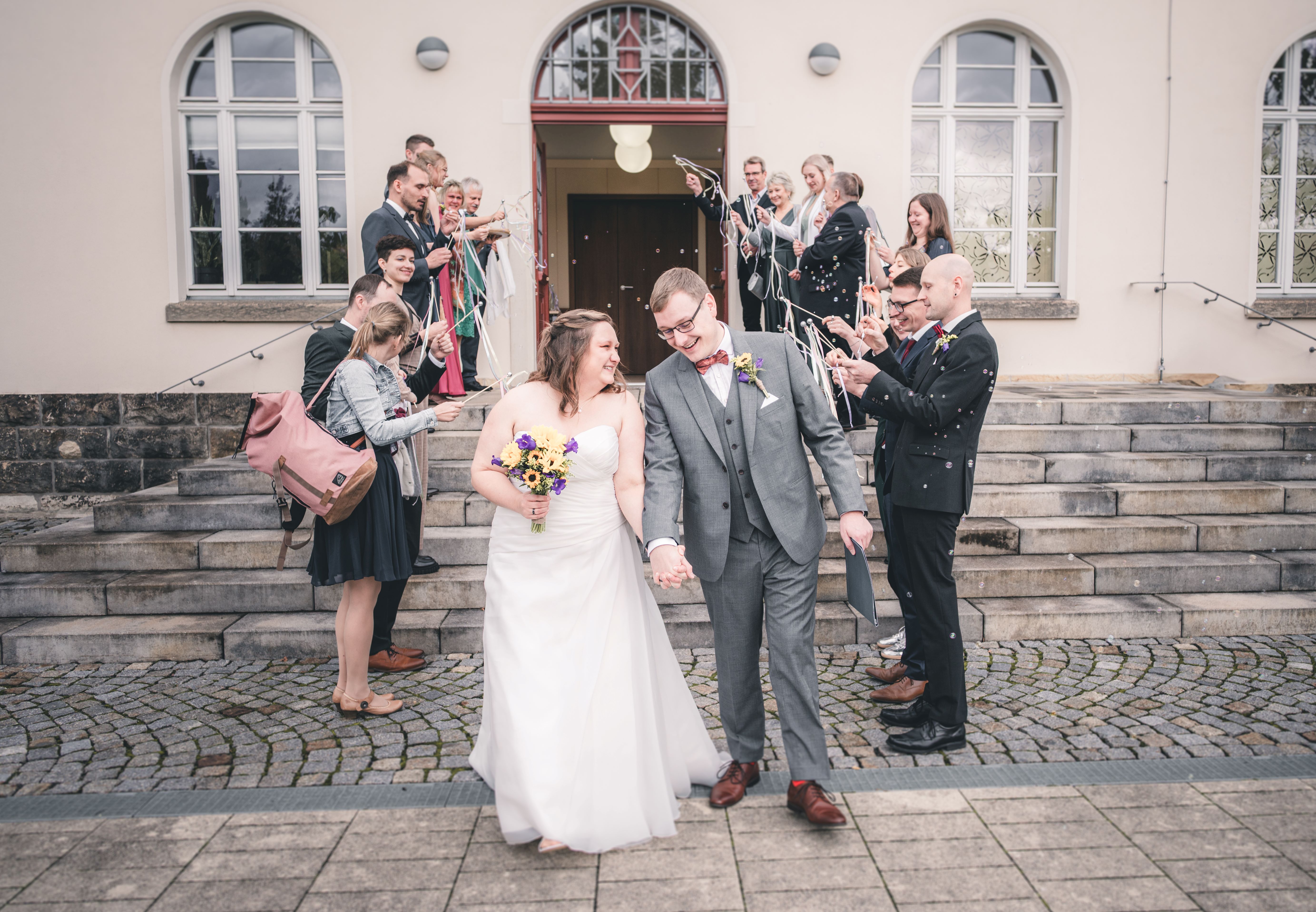 Das frisch verheiratete Brautpaar zieht glücklich lachend aus dem Standesamt Radebeul aus, während die Gäste Seifenblasen und Wedding Wands schwingen.