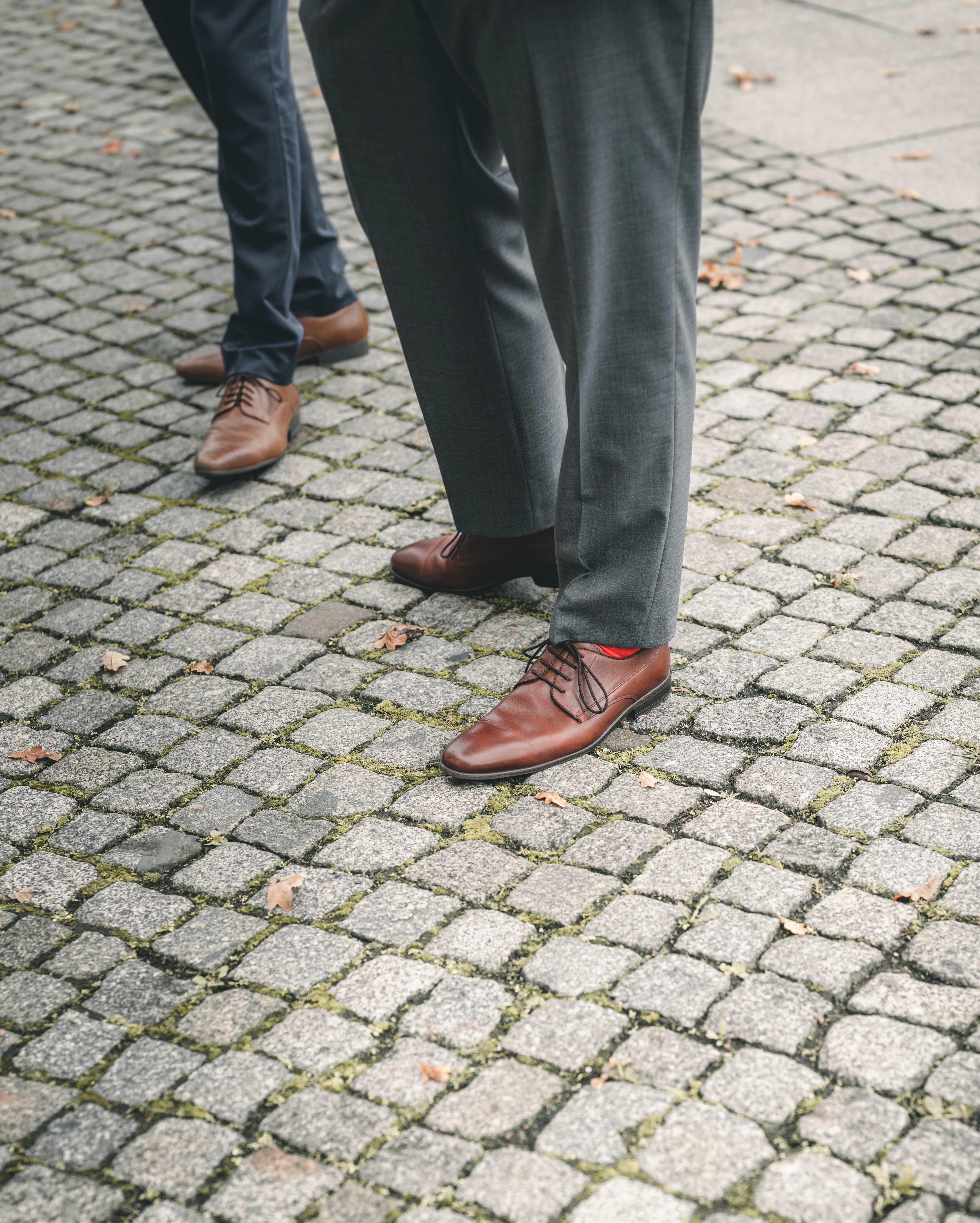 Eine detailreiche Nahaufnahme der braunen Lederschuhe des Bräutigams auf Pflastersteinen, bei der seine auffälligen roten Socken hervorblitzen.