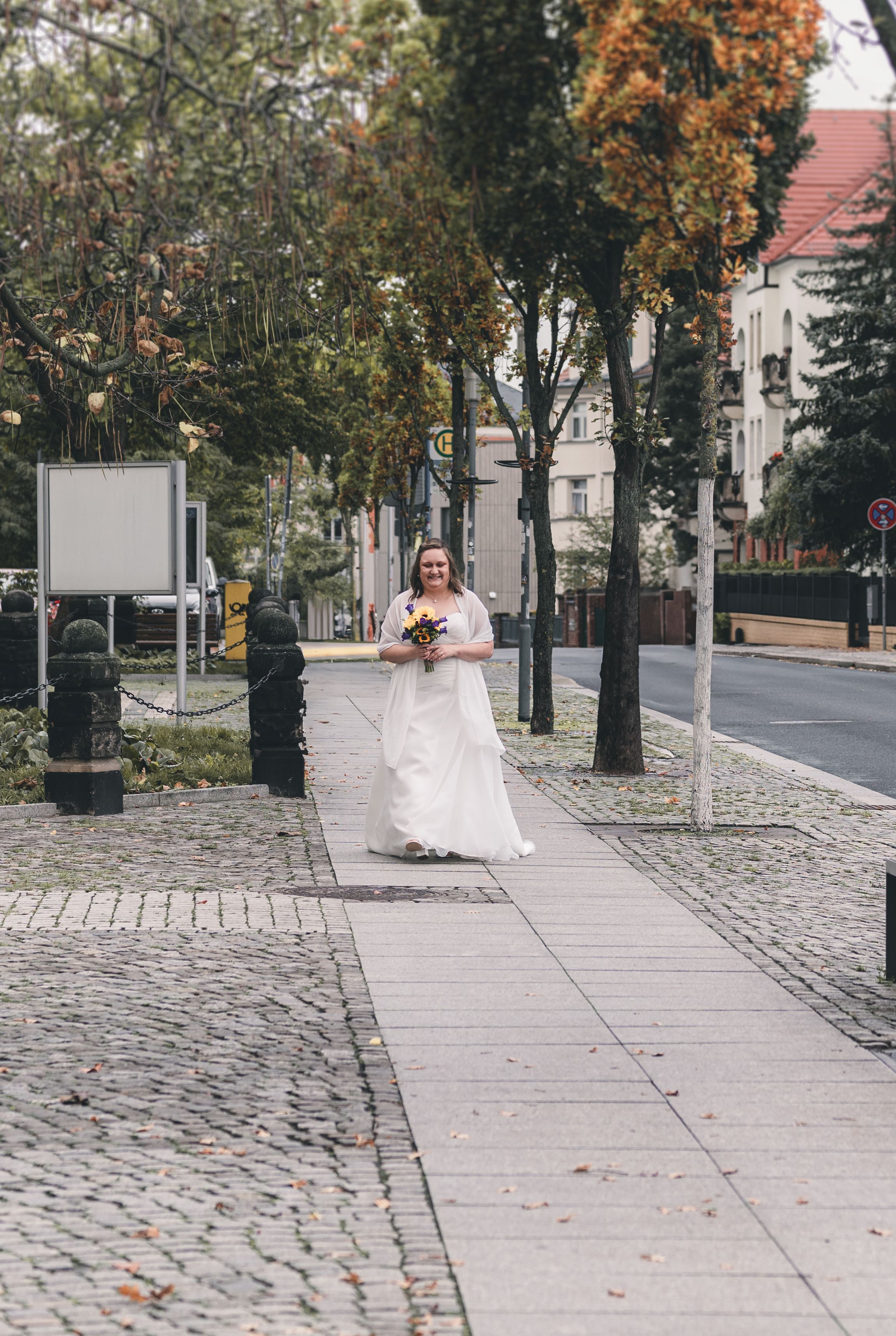 Die Braut läuft im weißen Hochzeitskleid mit einem farbenfrohen Sonnenblumenstrauß lächelnd den Gehweg zum Standesamt Radebeul entlang.