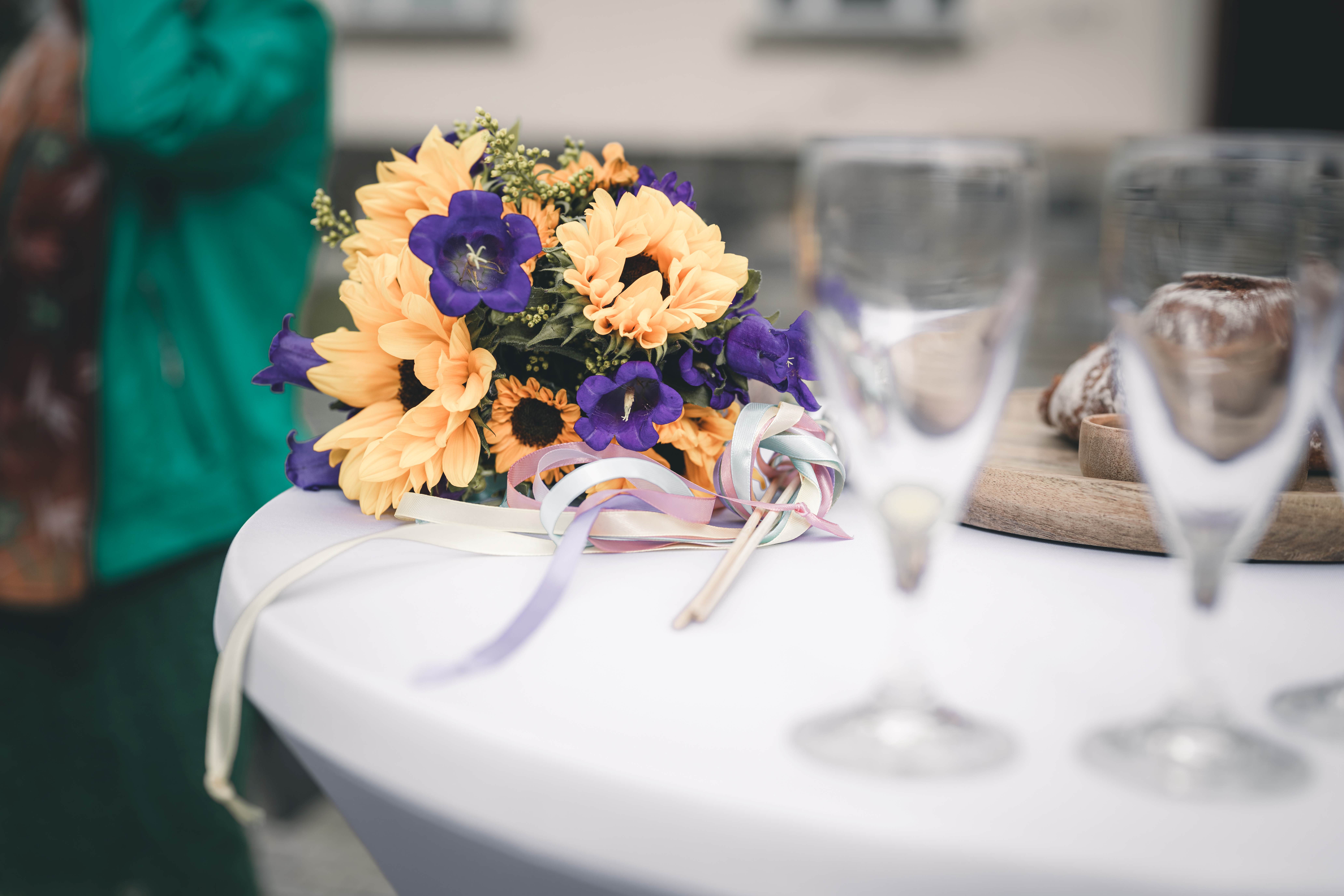 Ein herbstlicher Brautstrauß aus leuchtenden gelben Sonnenblumen und violetten Glockenblumen liegt neben Sektgläsern auf einem weißen Tisch.