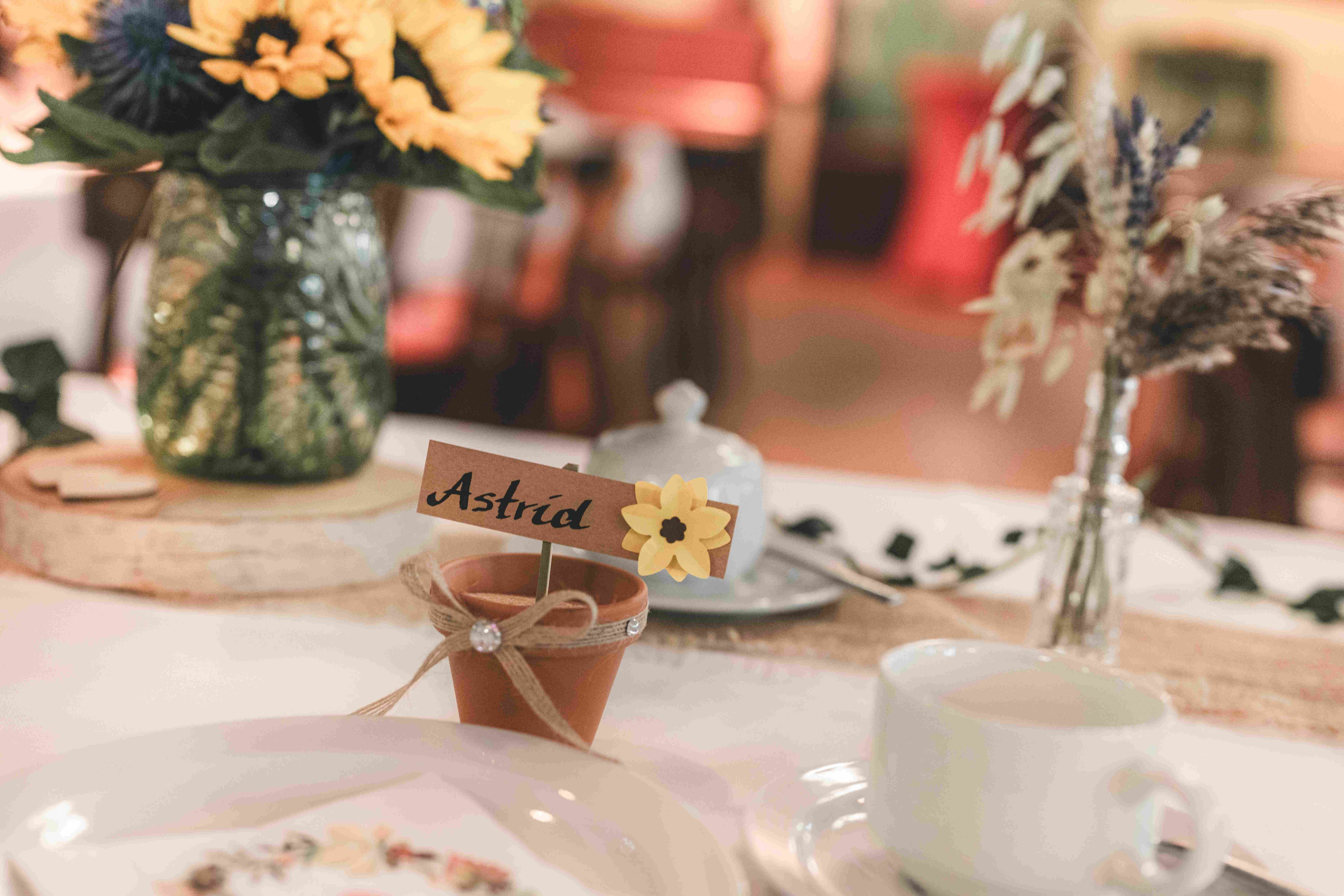 Ein liebevoll gestaltetes Namensschild mit der Aufschrift „Astrid“ steckt in einem kleinen Tontopf auf dem Hochzeitstisch neben einer Vase mit Sonnenblumen.