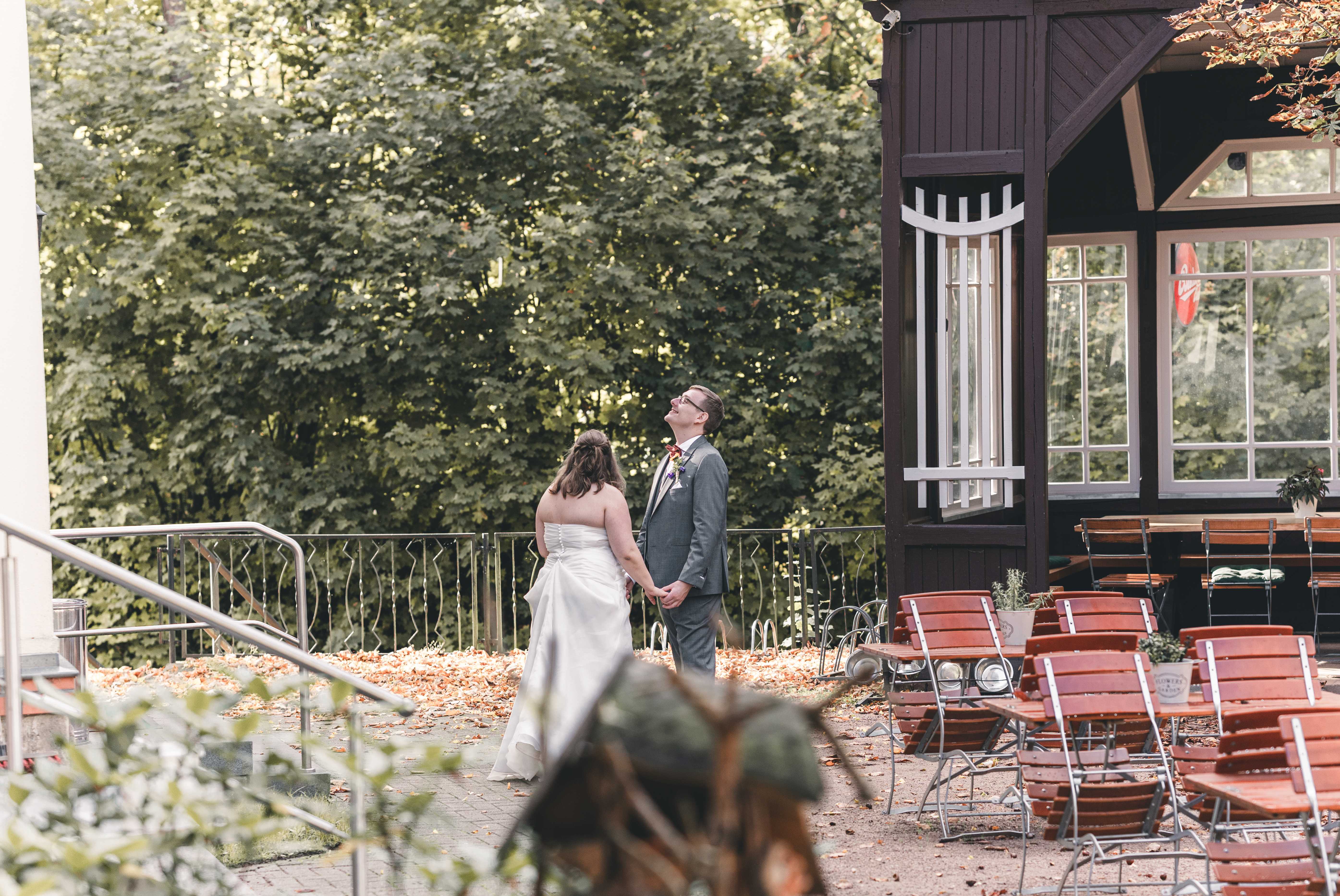 Das Brautpaar steht Hand in Hand auf der mit buntem Herbstlaub bedeckten Außenterrasse der Spitzgrundmühle und blickt gut gelaunt nach oben.