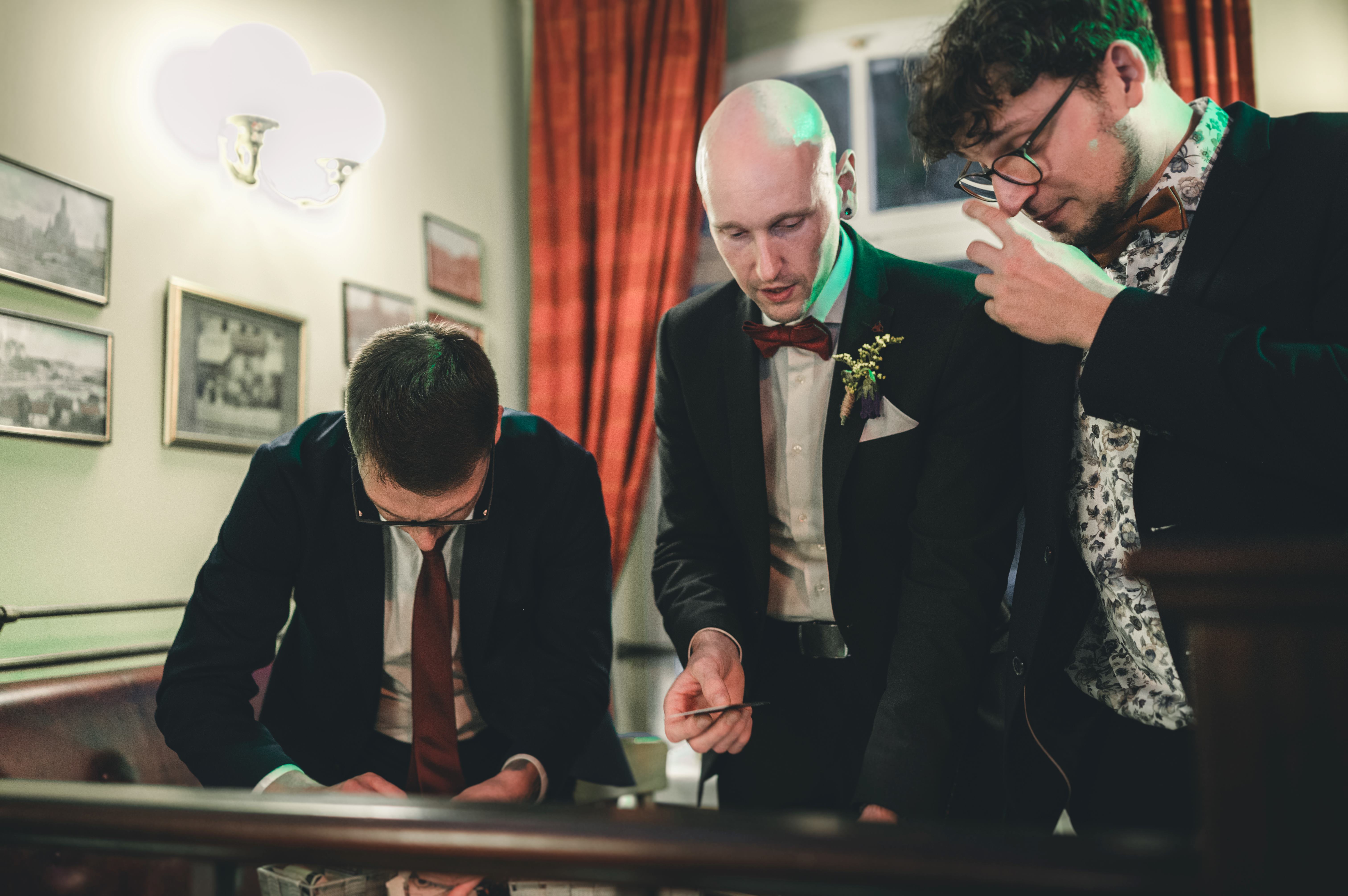 Drei Hochzeitsgäste in eleganten Anzügen begutachten gut gelaunt Spielkarten bei der Hochzeitsfeier in Coswig nahe Dresden.