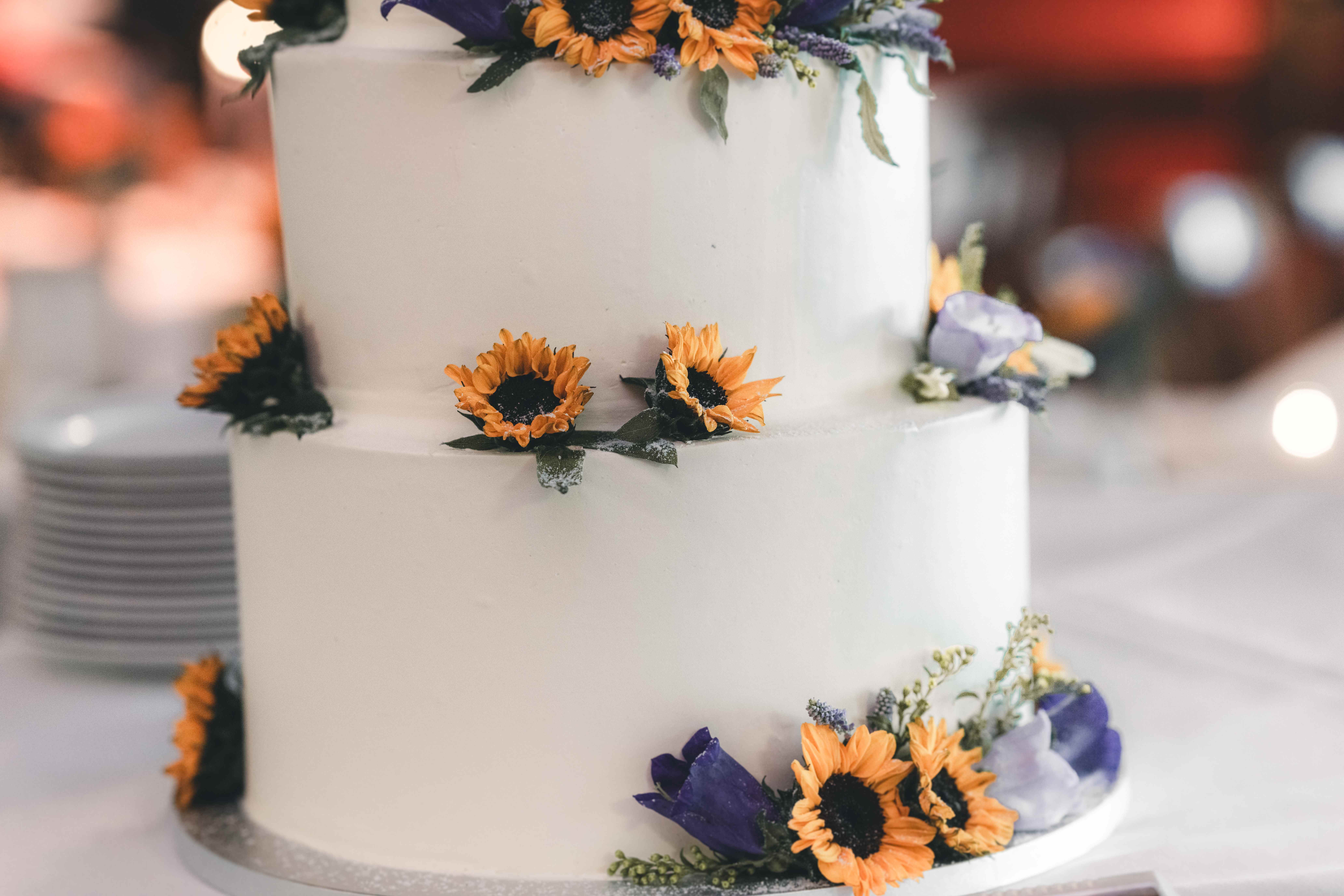 Nahaufnahme der weißen, dreistöckigen Hochzeitstorte, die kunstvoll mit frischen gelben Sonnenblumen und violetten Blüten verziert ist.