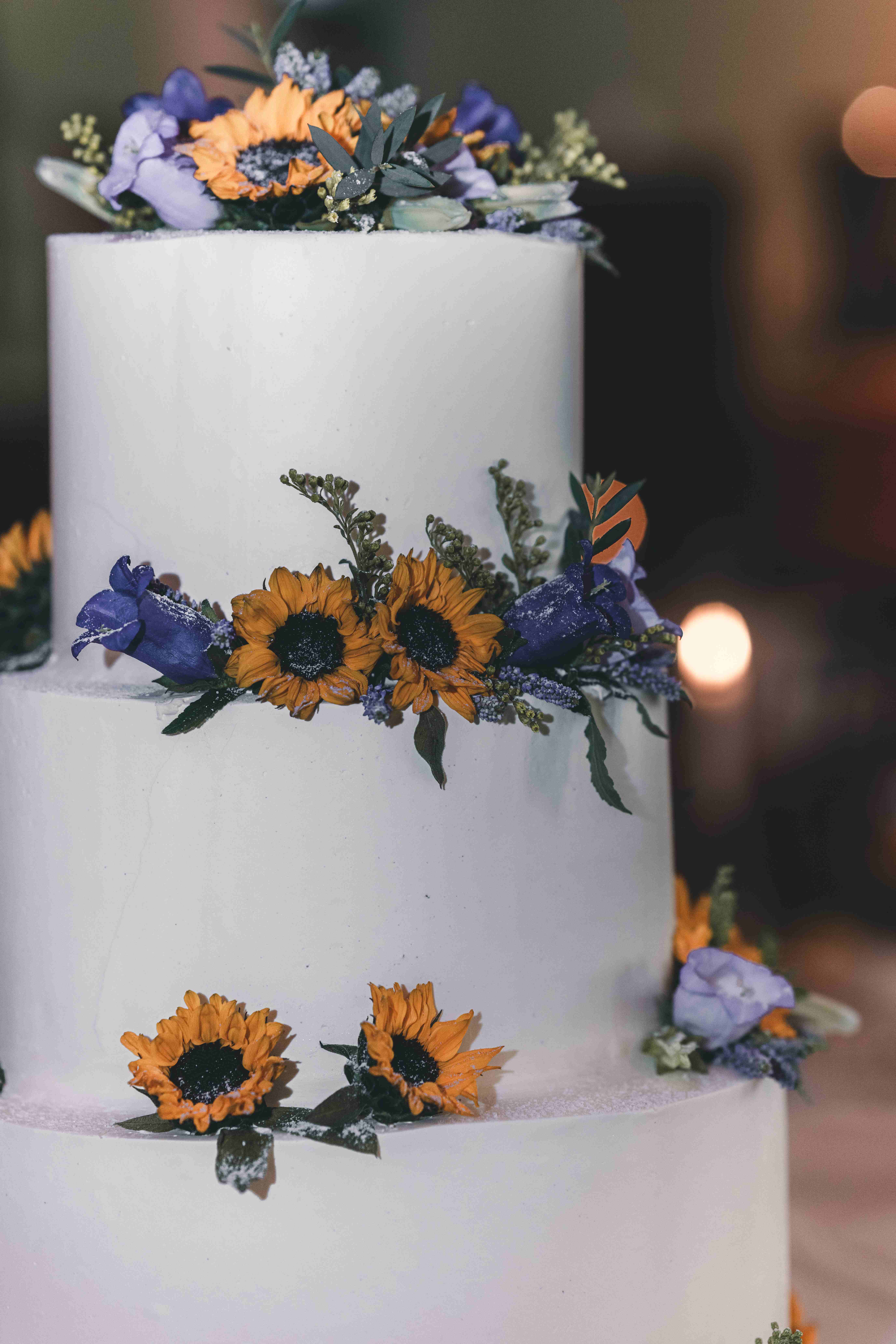 Das Brautpaar schneidet gemeinsam die dreistöckige, weiße Hochzeitstorte an, die liebevoll mit echten Sonnenblumen und lila Blüten verziert ist.