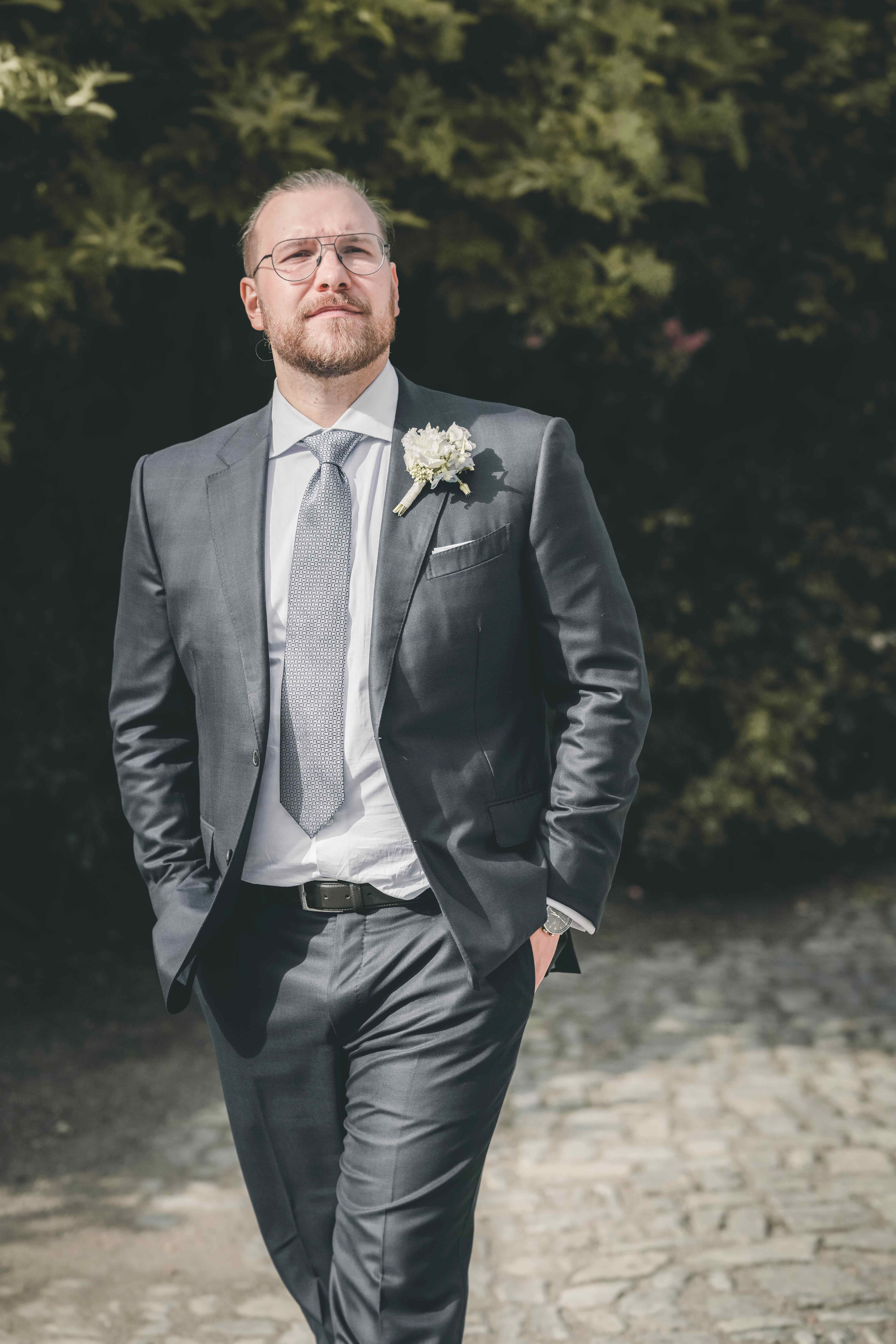 Porträt des Bräutigams mit Bart, Brille und gemusterter Krawatte, der die Hände in den Taschen hat und über das Kopfsteinpflaster geht.