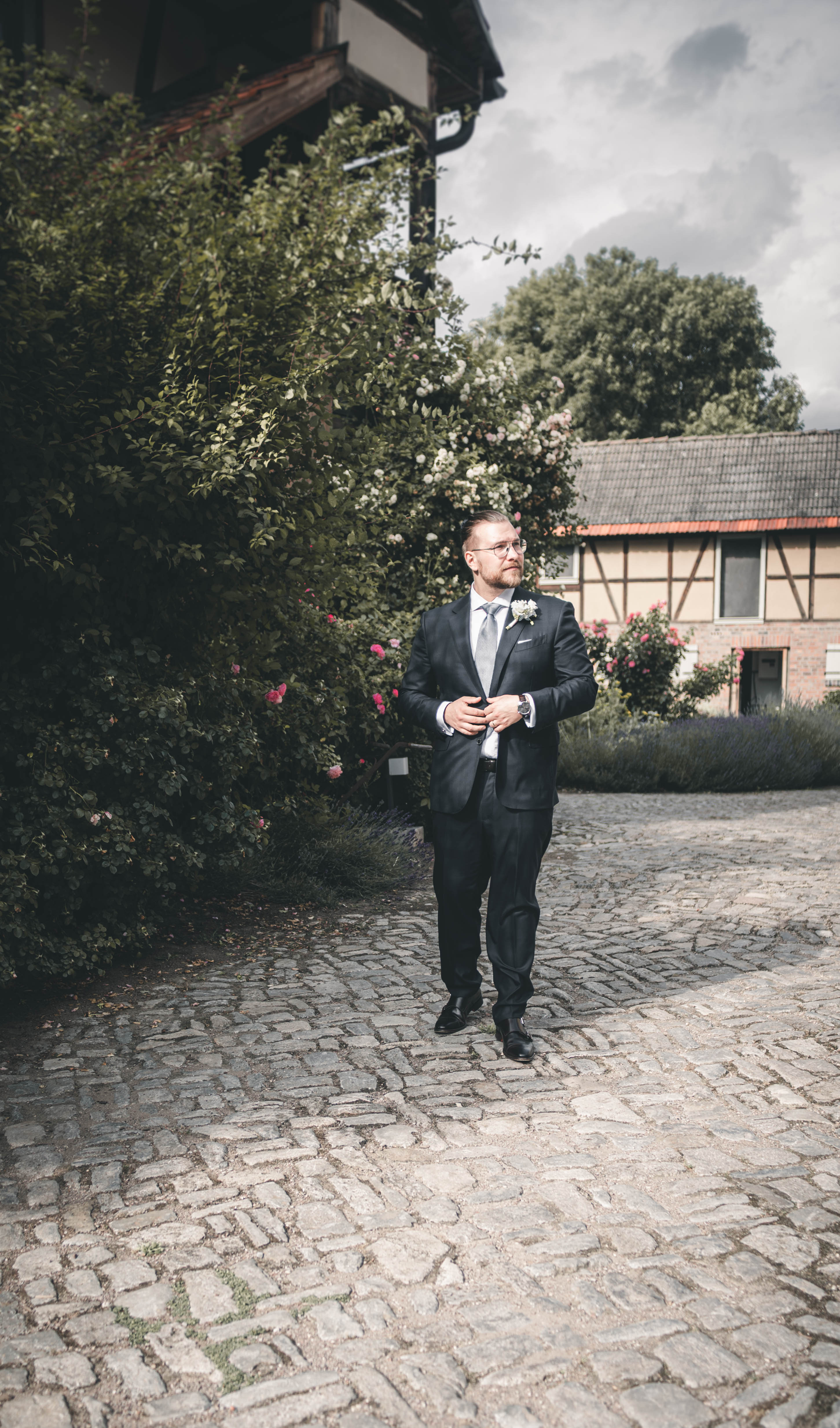 Der Bräutigam im dunklen Anzug schreitet mit Blick zur Seite über das Kopfsteinpflaster vorbei an Rosen und Fachwerk.