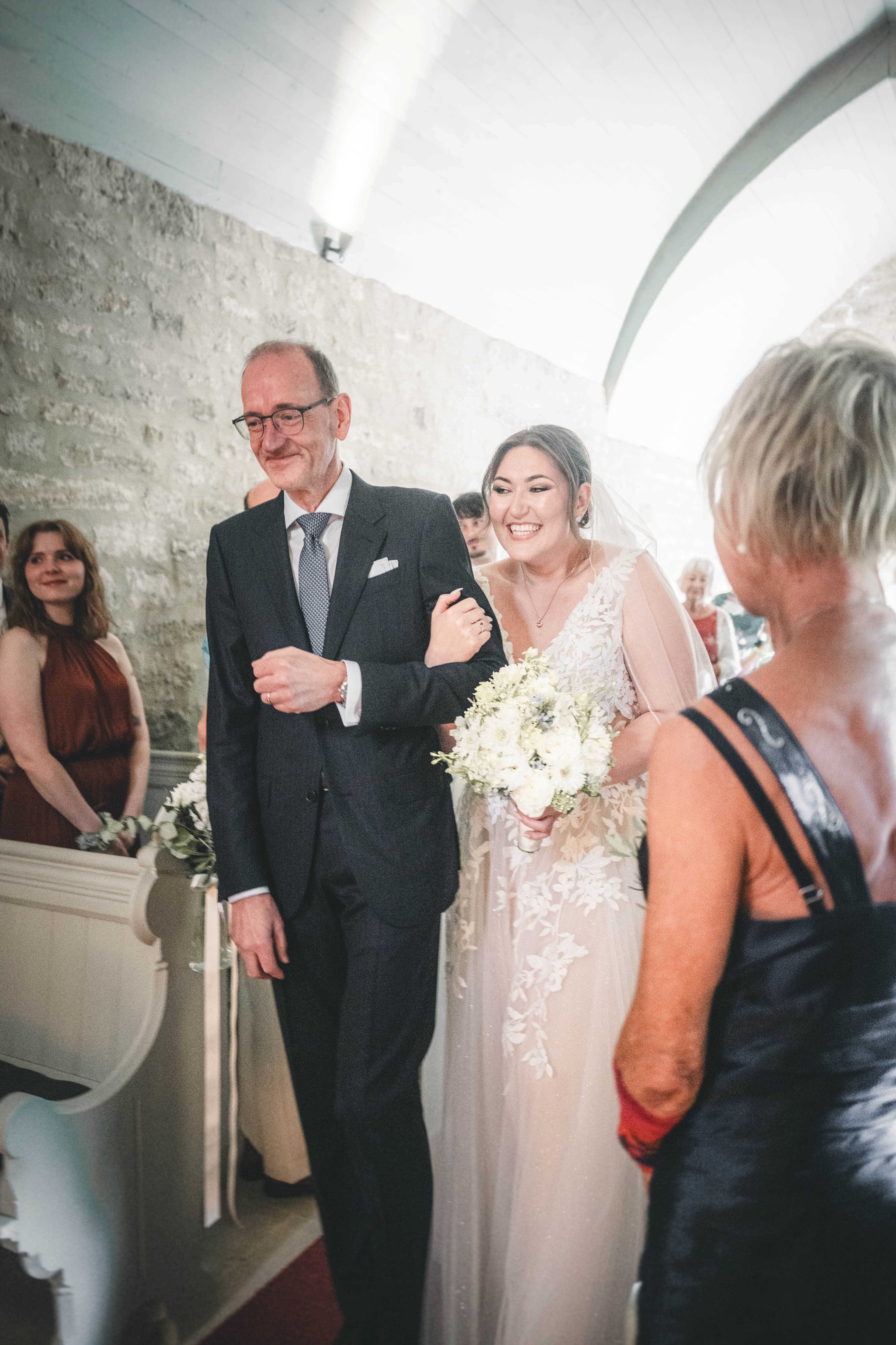 Die glücklich strahlende Braut im Spitzenkleid mit Brautstrauß zieht eingehakt bei ihrem Vater durch den Mittelgang der Kapelle im Magdalenengut Erfurt ein.