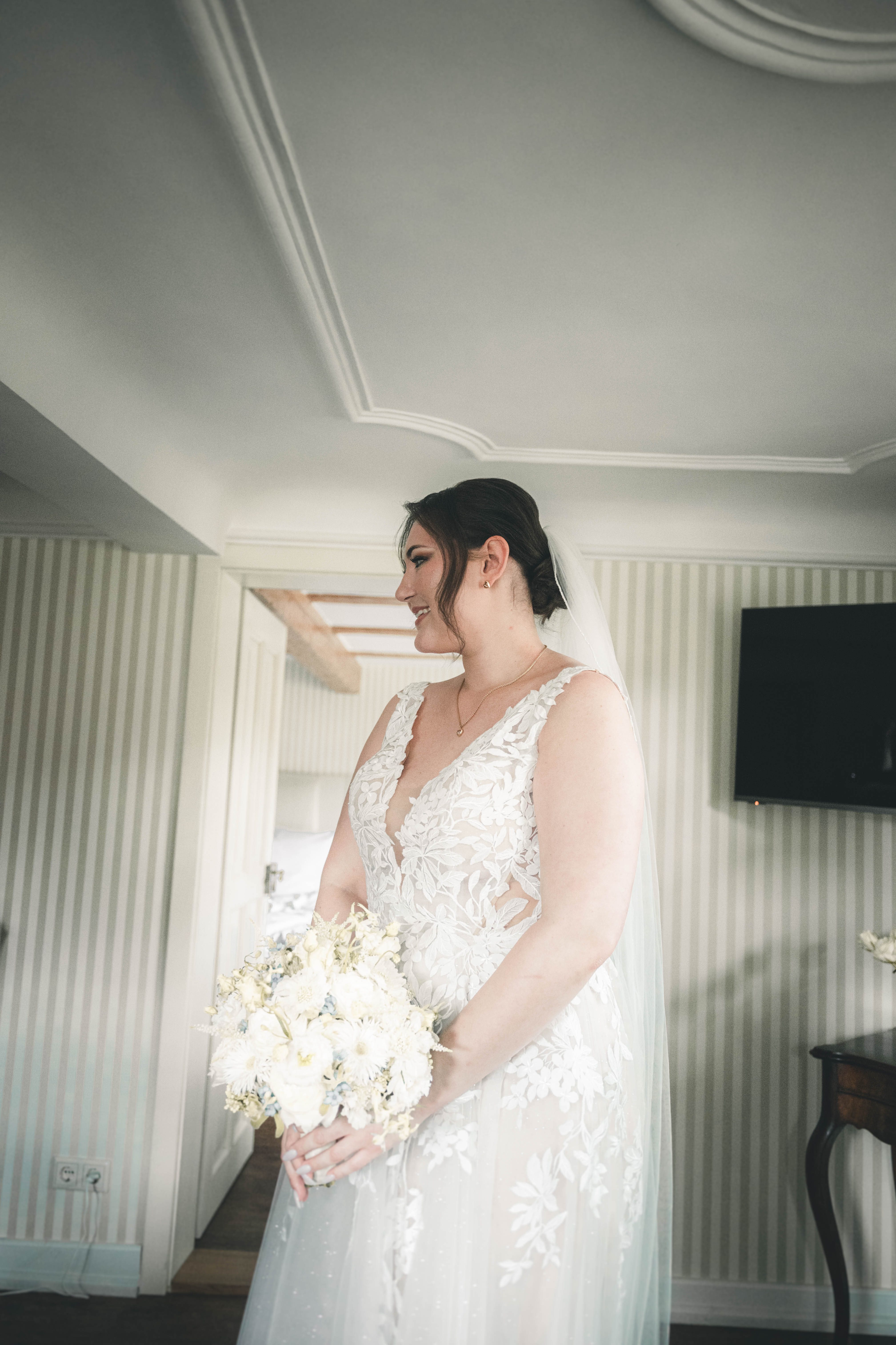 Die glückliche Braut im weißen Spitzenbrautkleid mit langem Tüllschleier und weißem Blumenstrauß steht im eleganten Raum des Magdalenenguts Erfurt.
