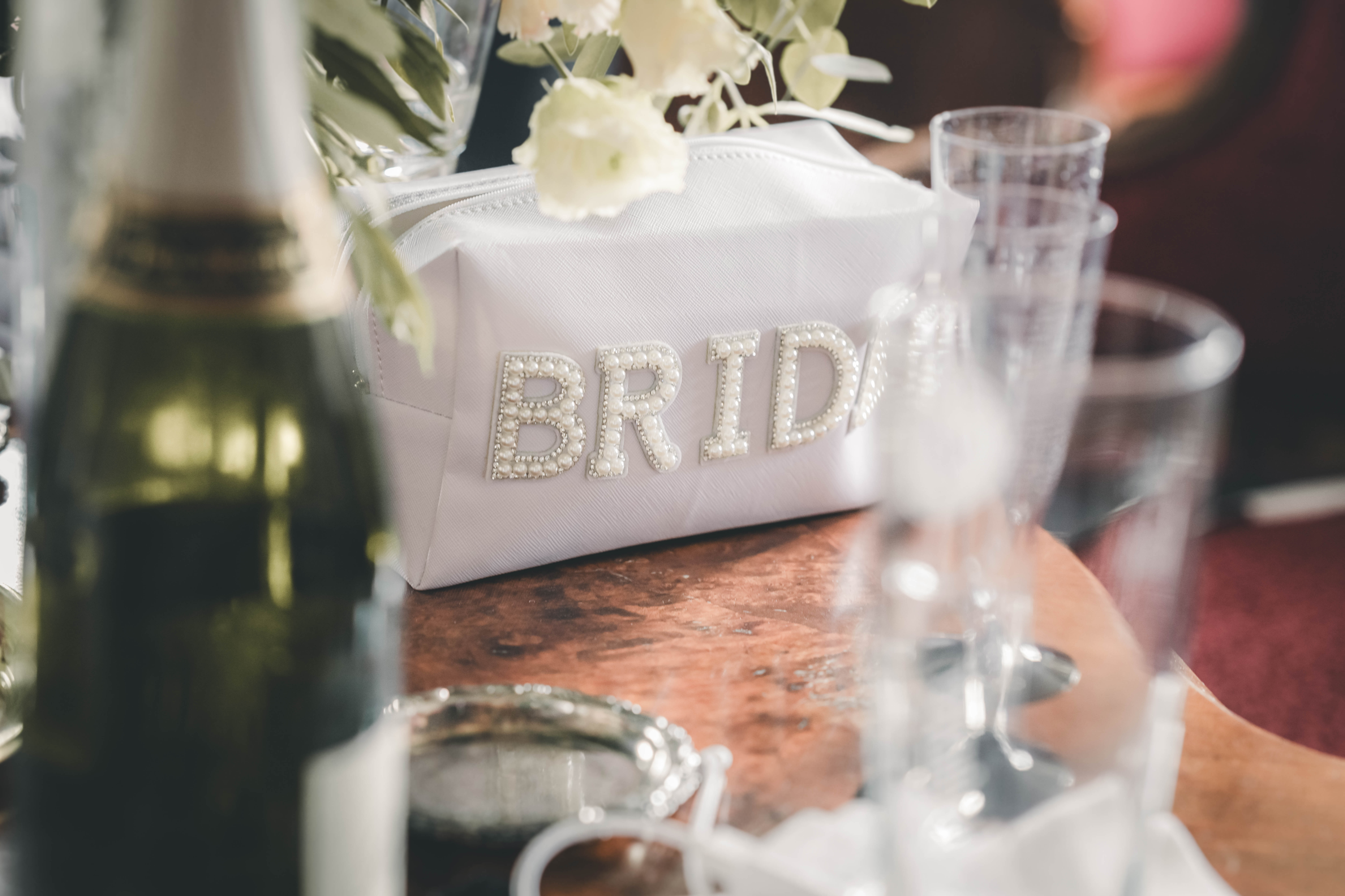 Weiße Make-up-Tasche mit aufgesticktem Perlen-Schriftzug „BRIDE“ neben Sektgläsern und Champagnerflasche auf einem Holztisch im Magdalenengut Erfurt.