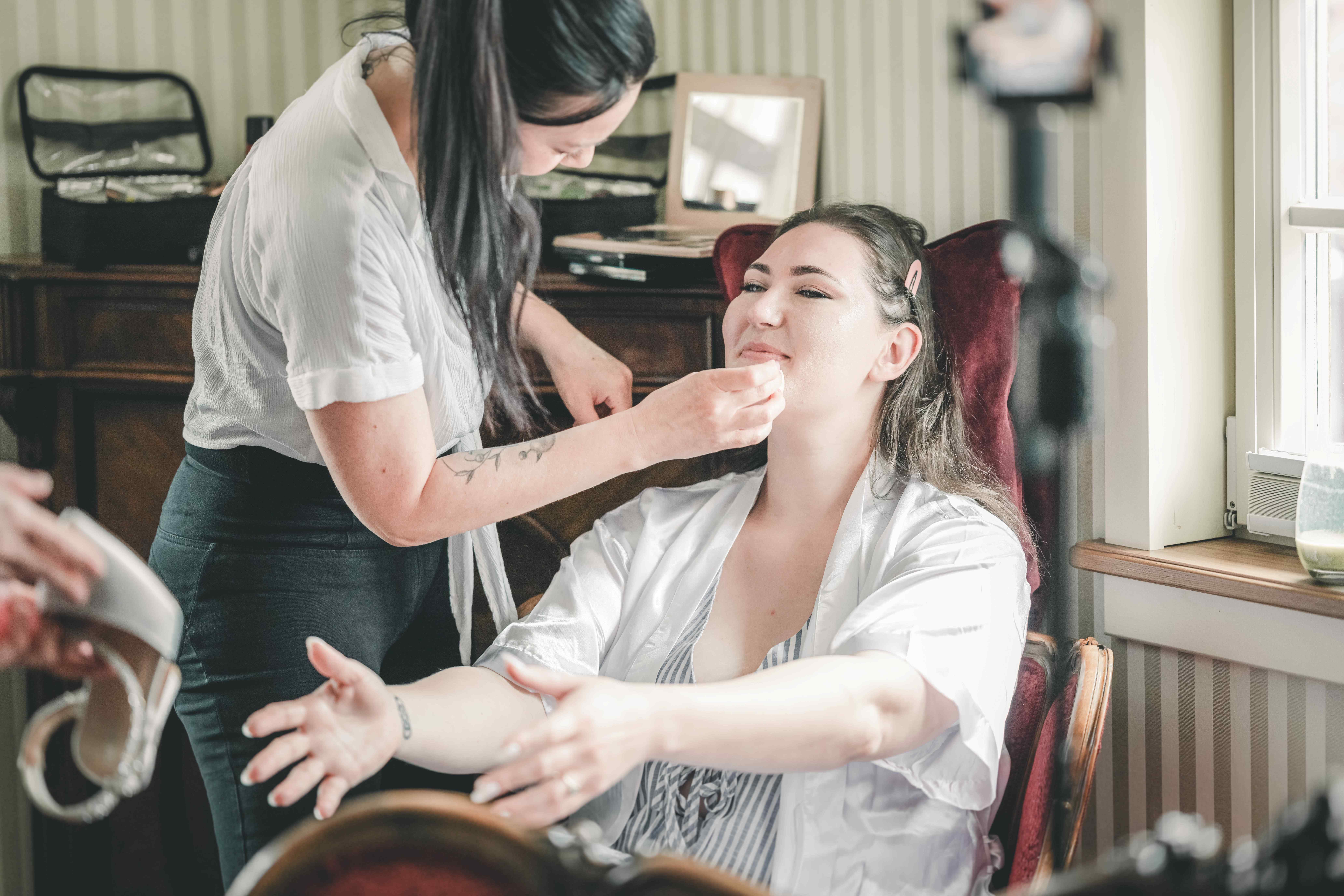 Die Braut im weißen Seidenmantel sitzt im Samtsessel und wird von der Stylistin für die Hochzeit im Magdalenengut Erfurt geschminkt.