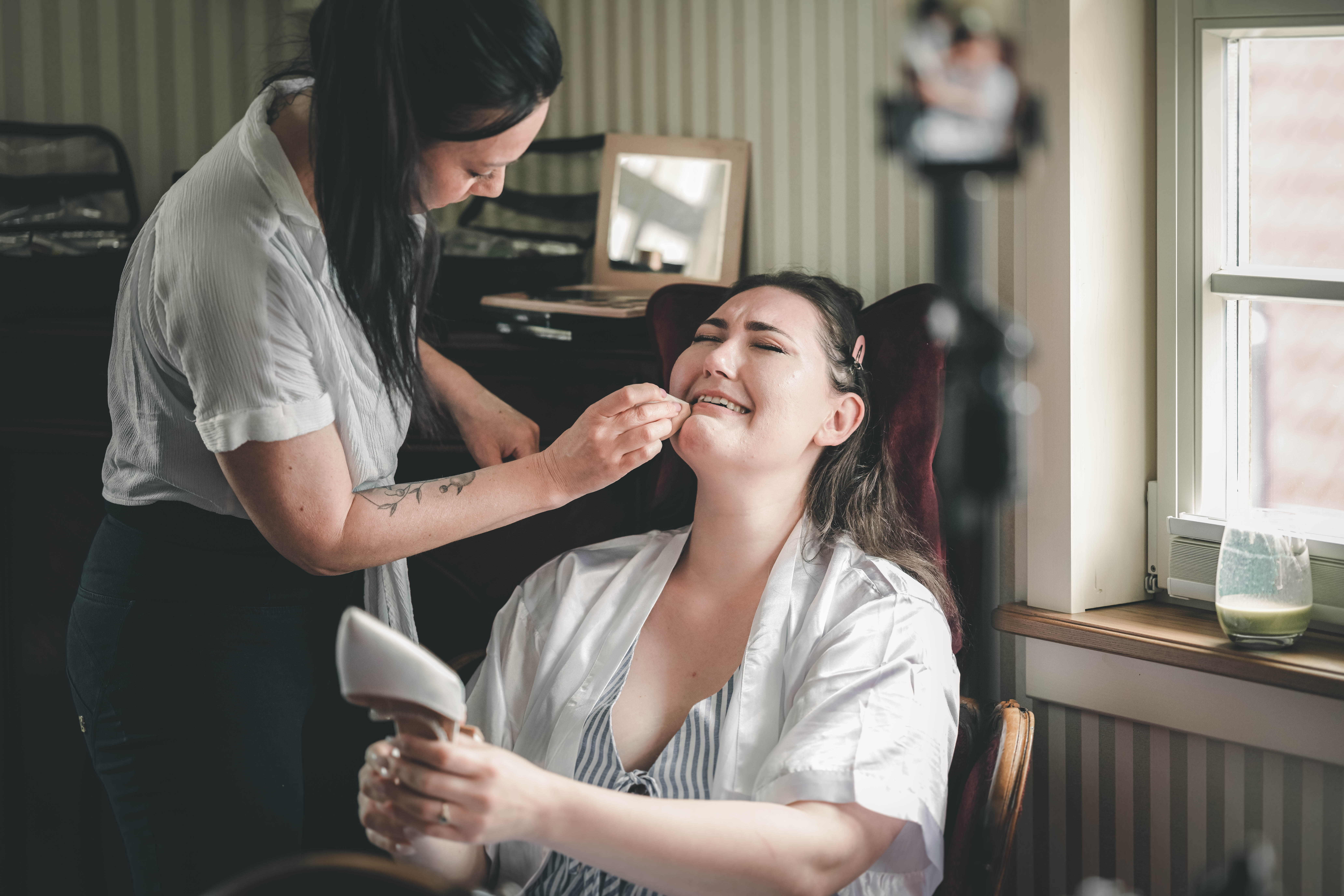 Eine Visagistin mit dunklen Haaren und einem weißen Hemd trägt der Braut vorsichtig Lippenstift auf. Die Braut hat die Augen geschlossen und sitzt in einem roten Sessel.