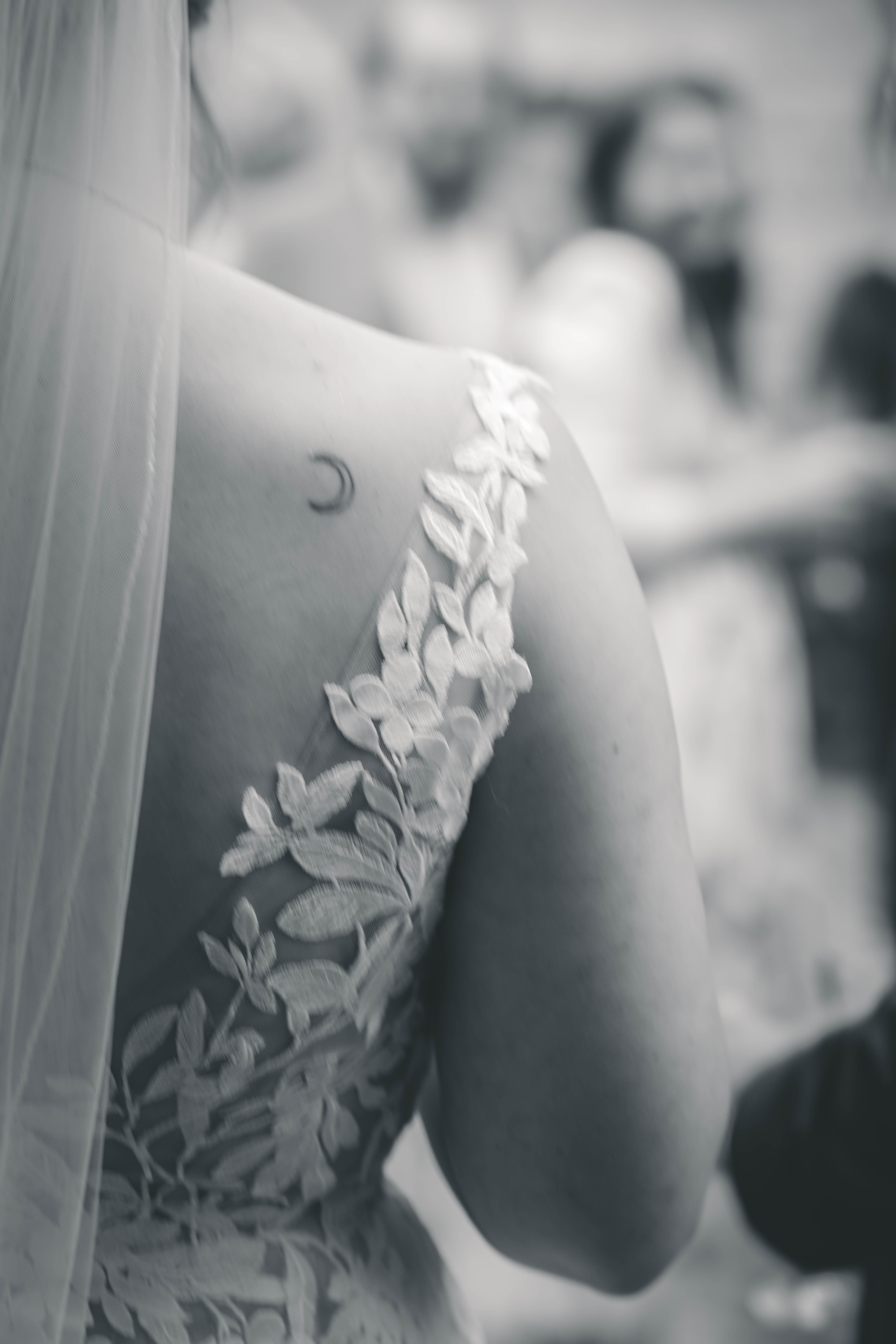 Schwarz-Weiß-Aufnahme: Detail des floralen Spitzenkleids und Tüllschleiers mit einem zarten Halbmond-Tattoo auf der Schulter der Braut.