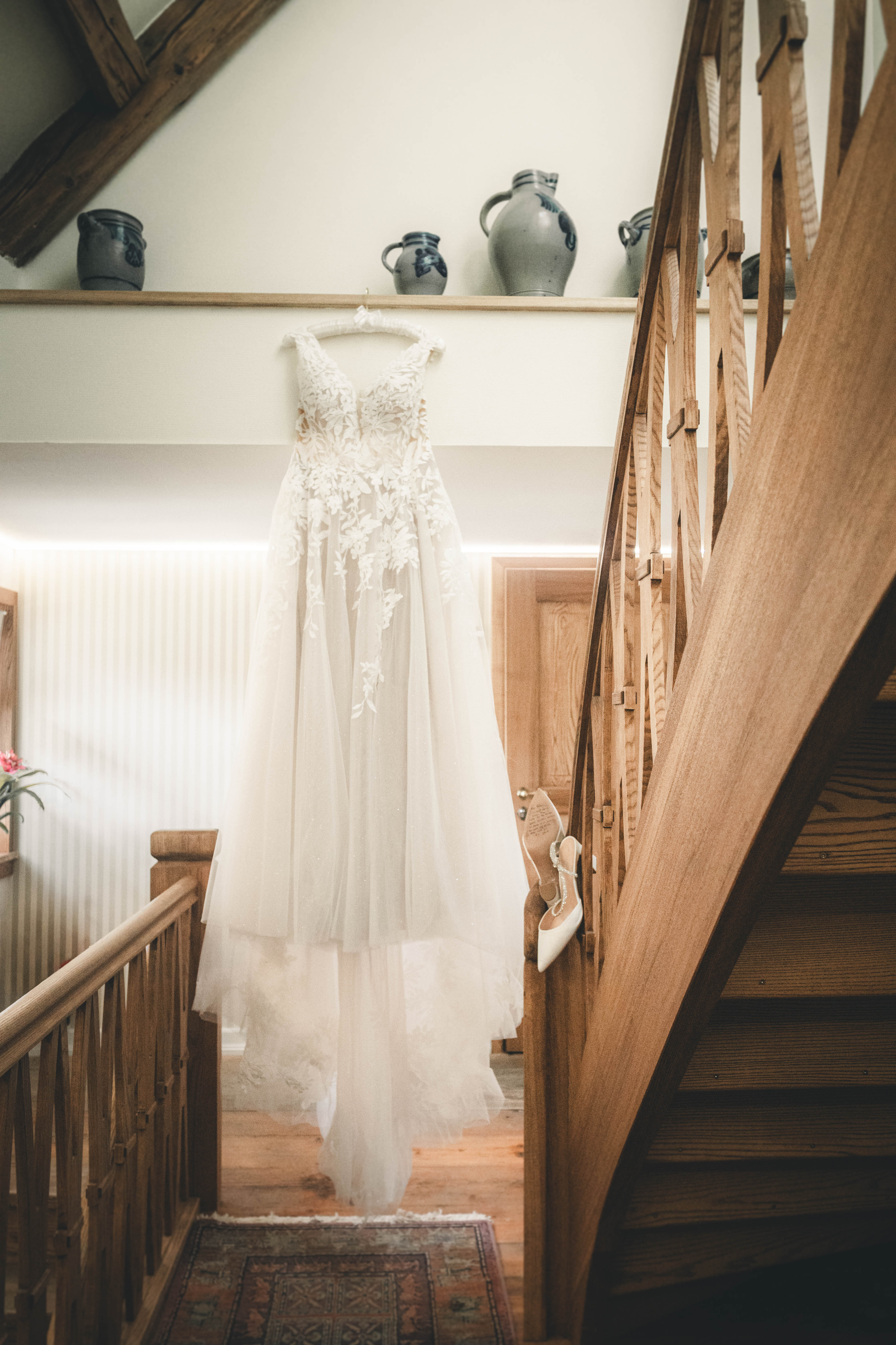 Ein bodenlanges Tüll-Brautkleid mit floralen Spitzenverzierungen hängt neben funkelnden Brautschuhen an der Holztreppe im Magdalenengut Erfurt.