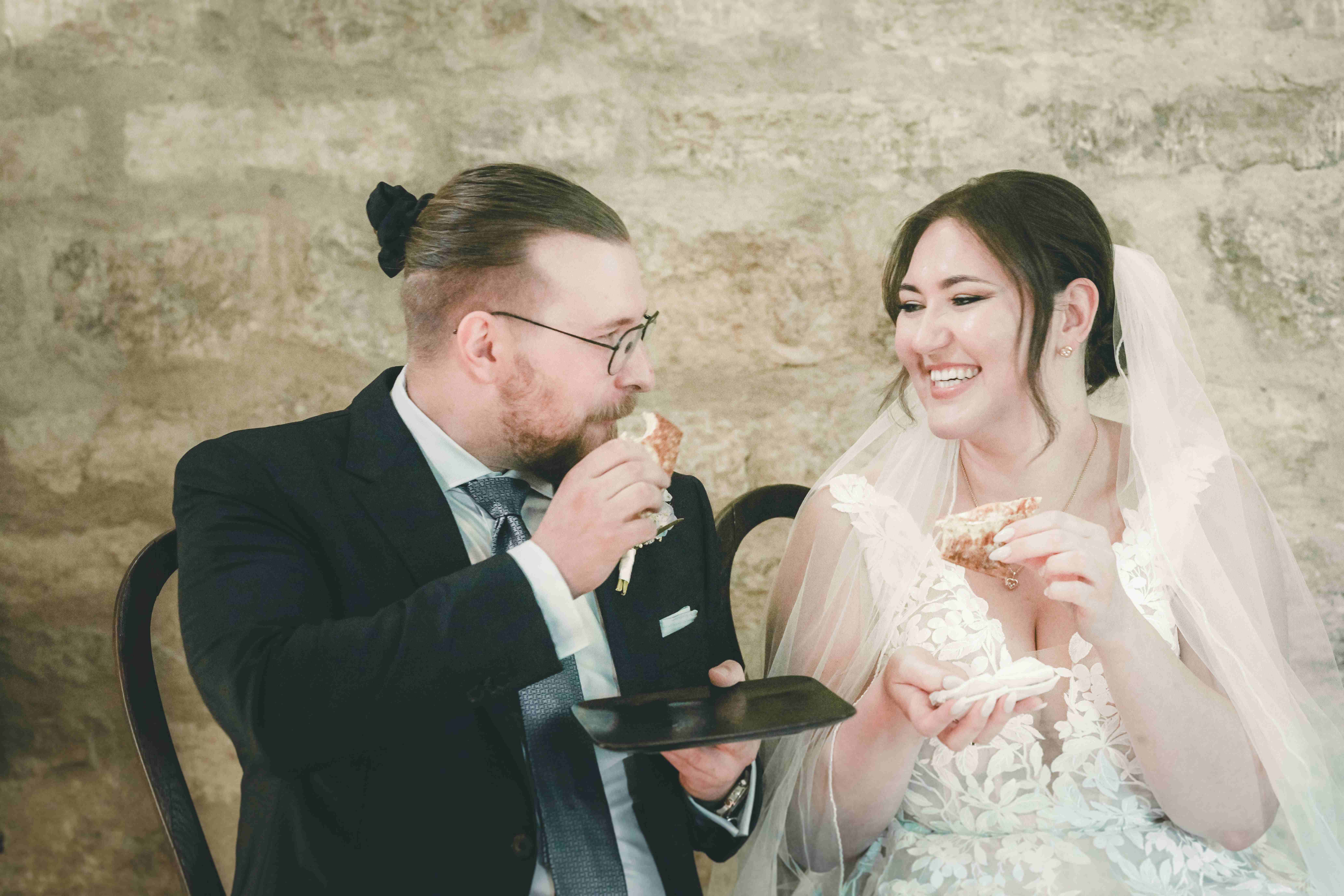 Braut und Bräutigam essen schmunzelnd ein Stück Brot während des Hochzeitsrituals vor der Natursteinkulisse im Magdalenengut Erfurt.