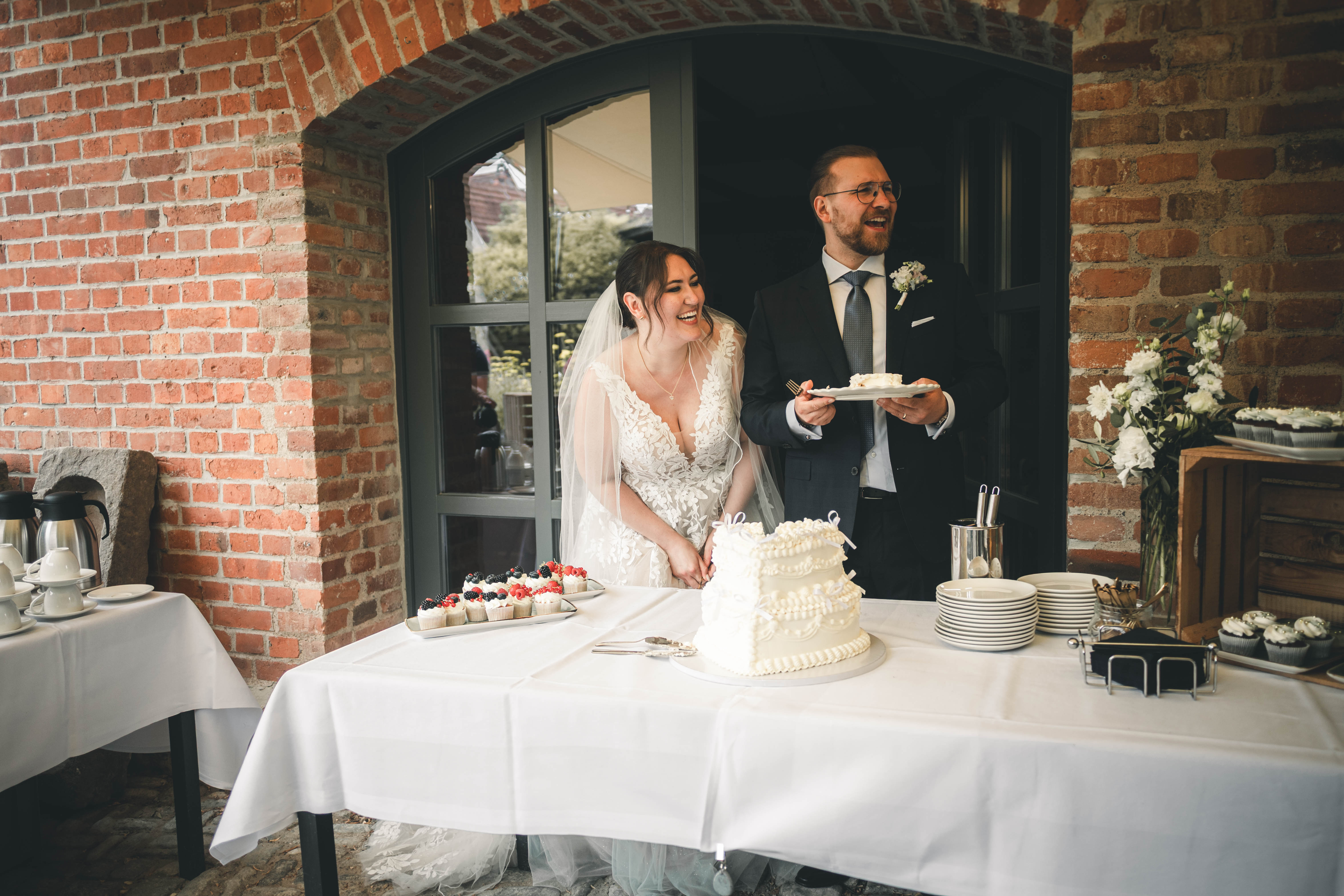 Die Braut im Spitzenkleid und der Bräutigam im dunklen Anzug lachen ausgelassen mit dem ersten Stück Hochzeitstorte im Magdalenengut Erfurt.