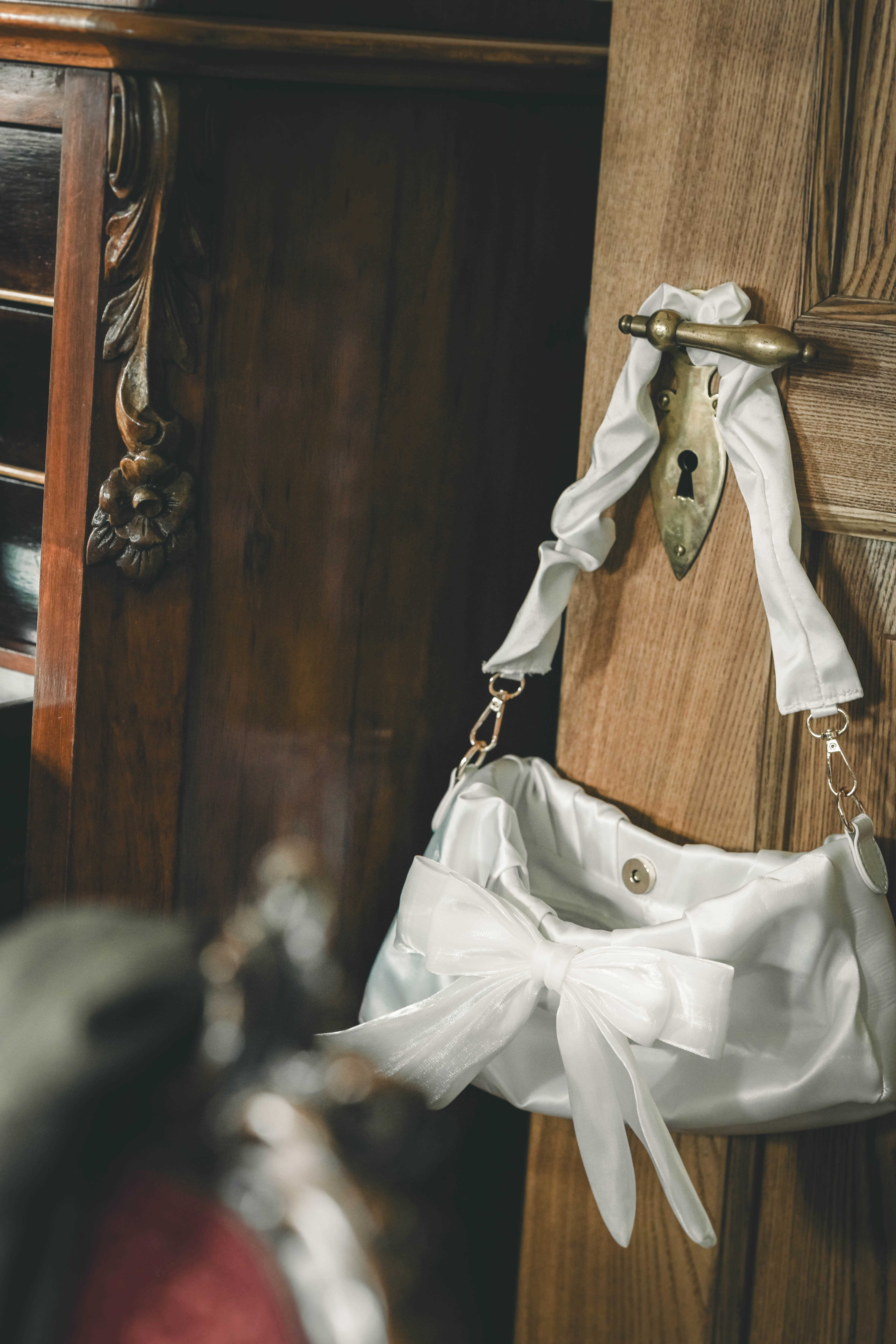 Weiße Brauttasche aus glänzendem Satin mit Schleifendetail hängt an der Messingklinke einer antiken Holztür im Magdalenengut Erfurt.