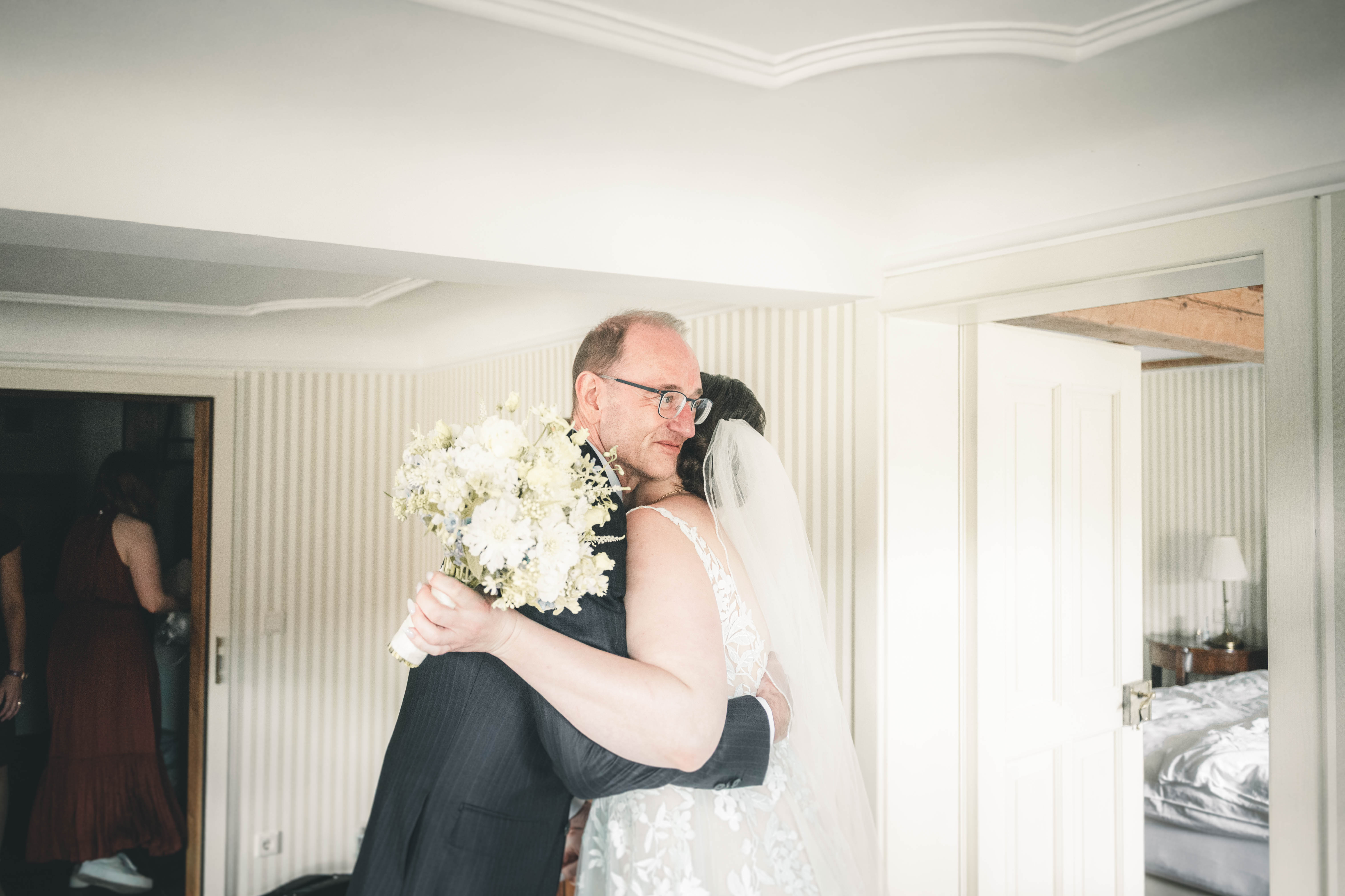 Der gerührte Vater umarmt seine Tochter im Brautkleid mit Schleier und weißem Blumenstrauß fest im Salon des Magdalenenguts Erfurt.
