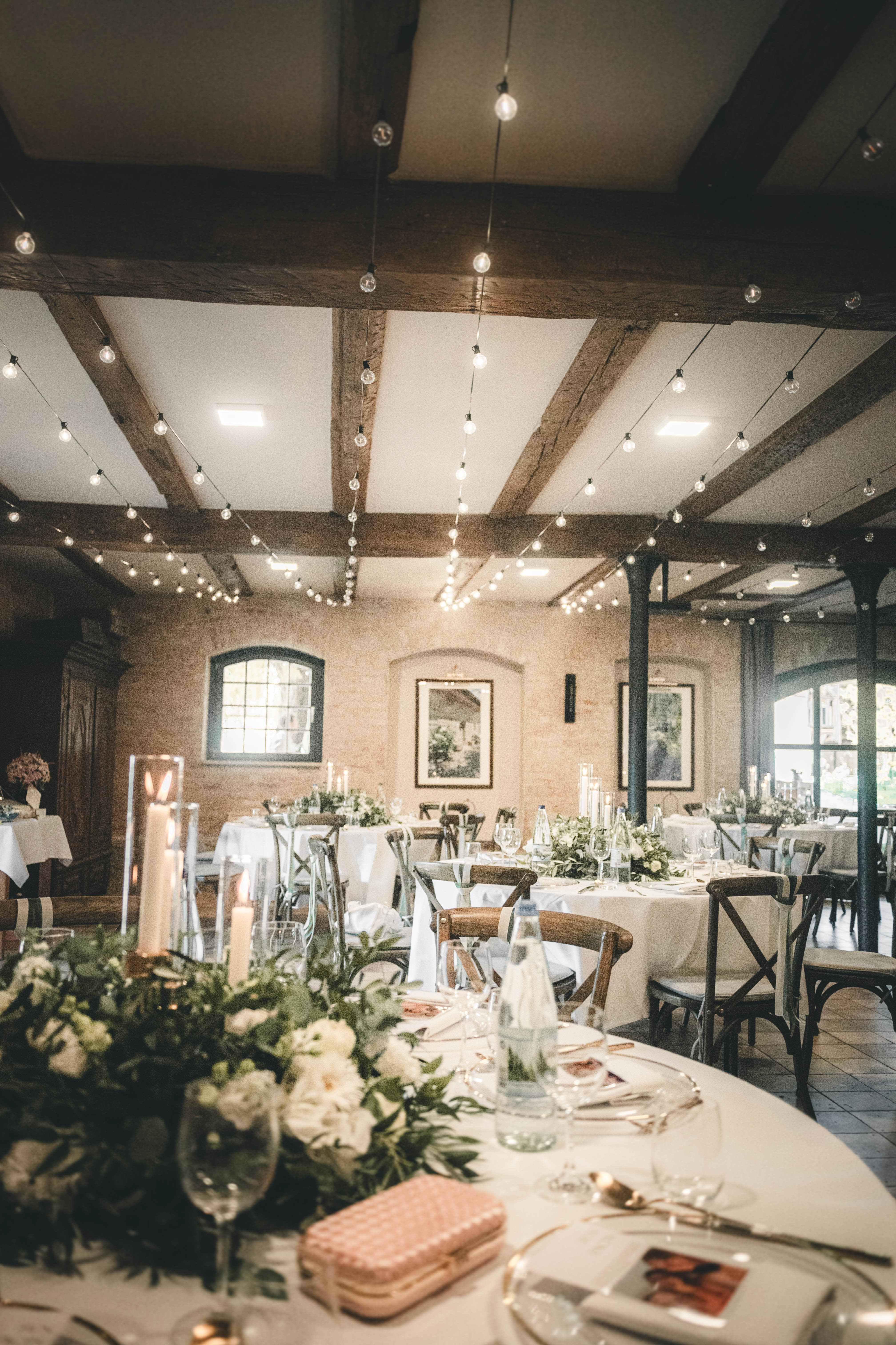 Warmer Festsaal im Magdalenengut Erfurt mit runden Tischen, Kerzenleuchtern, floralen Gestecken und hängenden Lichterketten.
