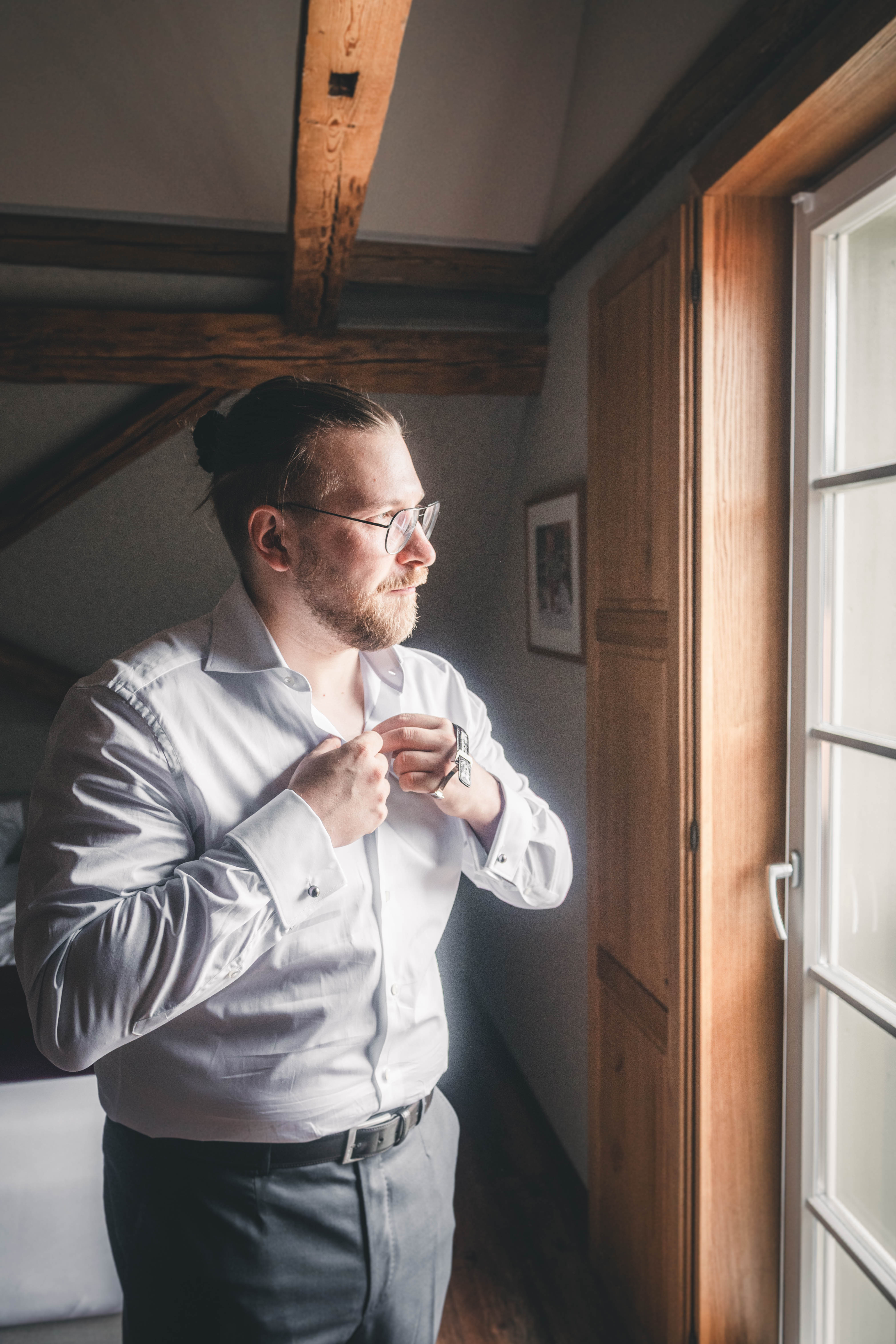 Der Bräutigam im weißen Hemd schließt seinen Kragen am lichtdurchfluteten Fenster im rustikalen Dachzimmer des Magdalenenguts Erfurt.