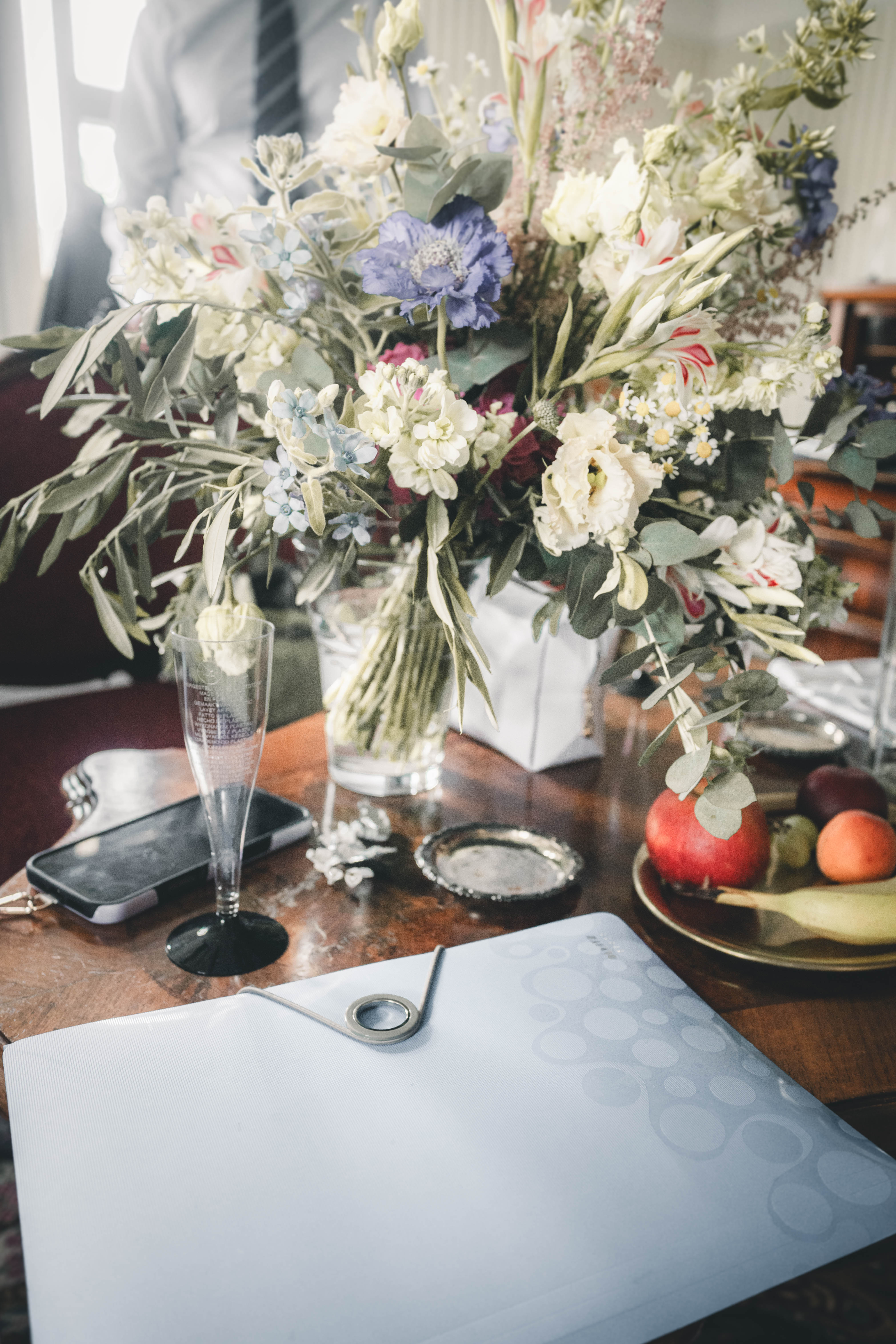 Stillleben auf einem dunklen Holztisch mit einem großen Wildblumenstrauß in einer Vase, einem silbernen Laptop, einem Sektglas, einem Handy und einem Teller mit Früchten.