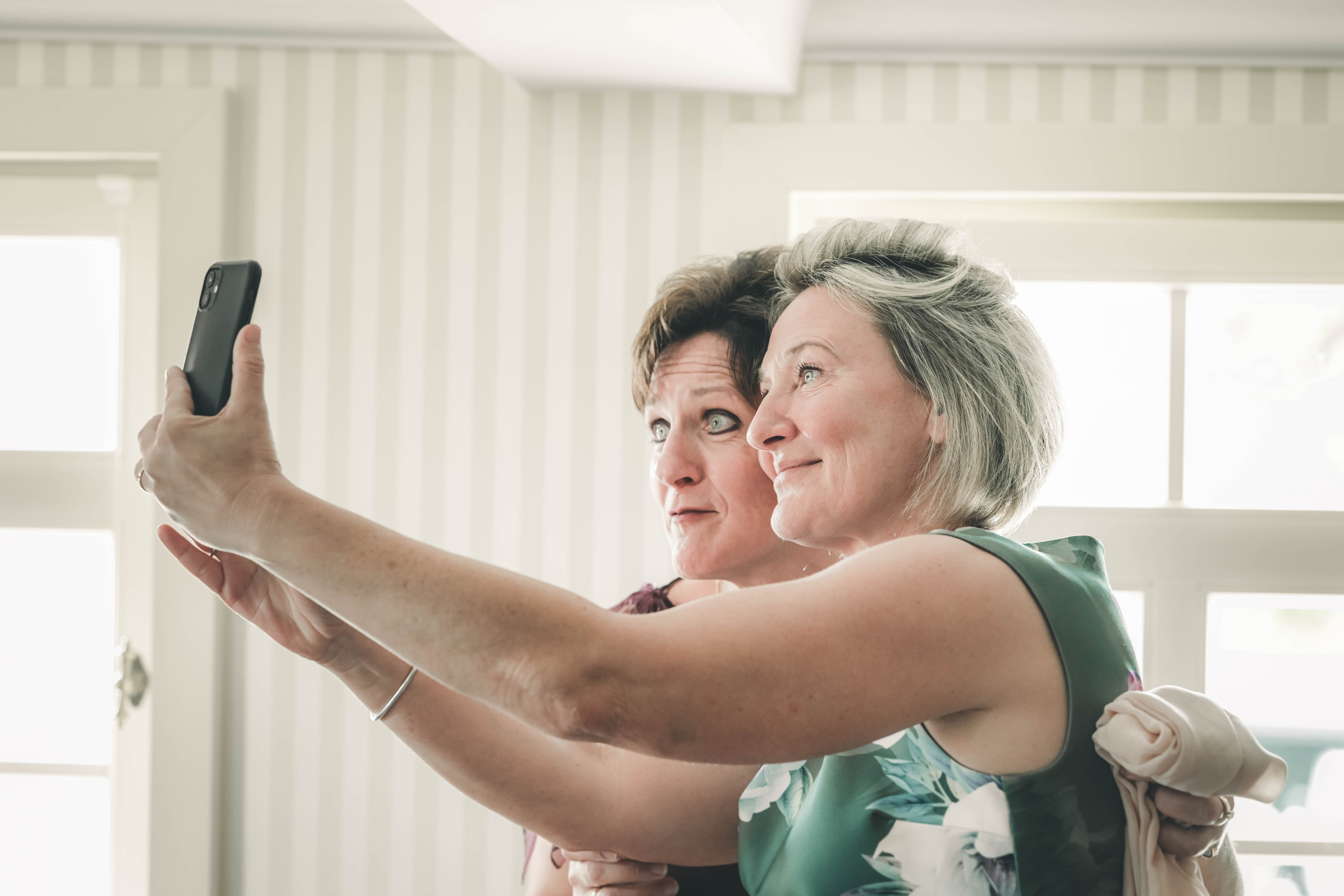 Zwei festlich gekleidete Frauen lächeln gemeinsam für ein Erinnerungs-Selfie mit dem Smartphone während des Getting Ready im Magdalenengut Erfurt.
