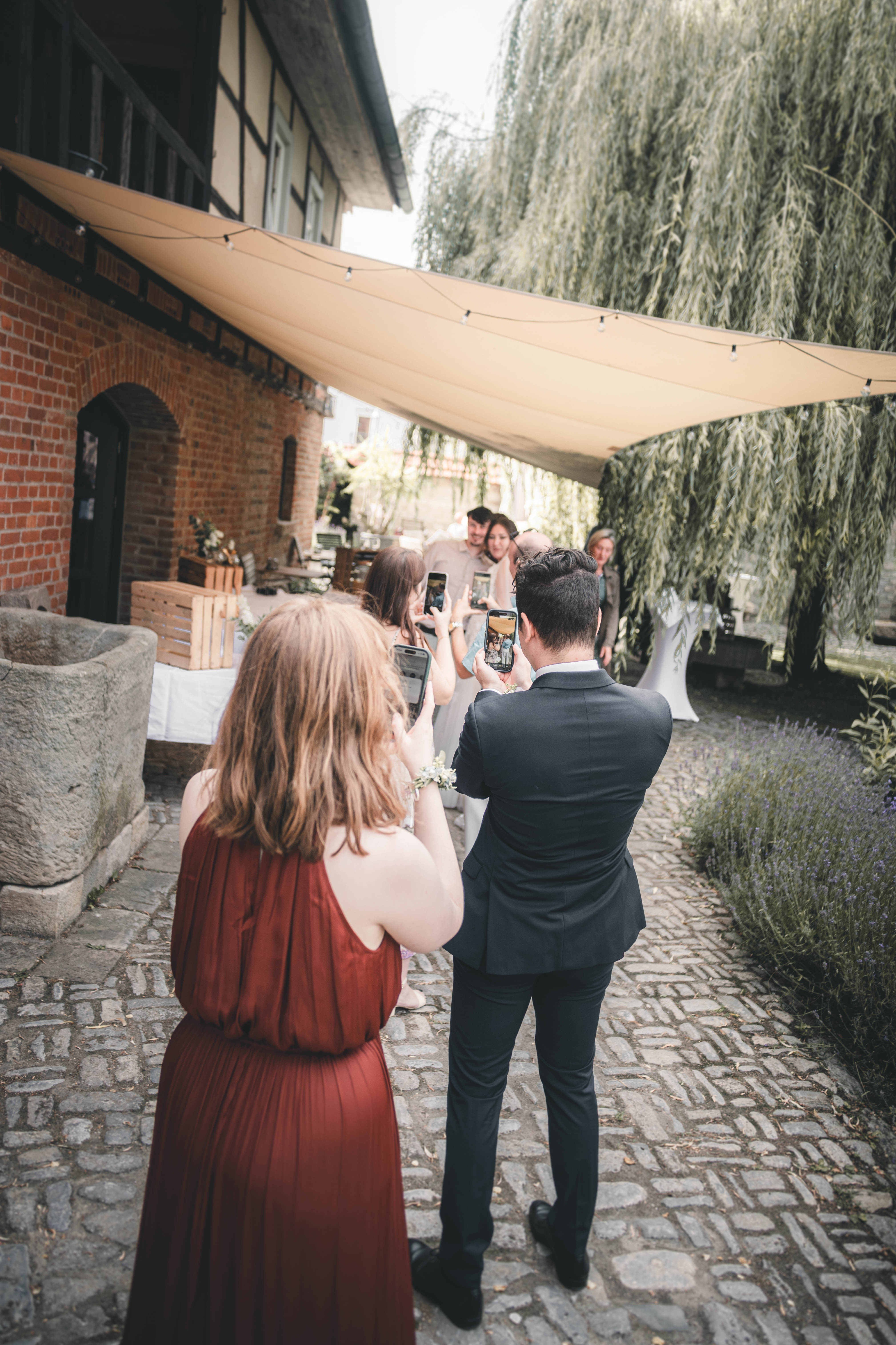 Gäste stehen auf dem Kopfsteinpflaster unter dem Sonnensegel und machen Fotos mit ihren Smartphones im Innenhof des Magdalenenguts Erfurt.