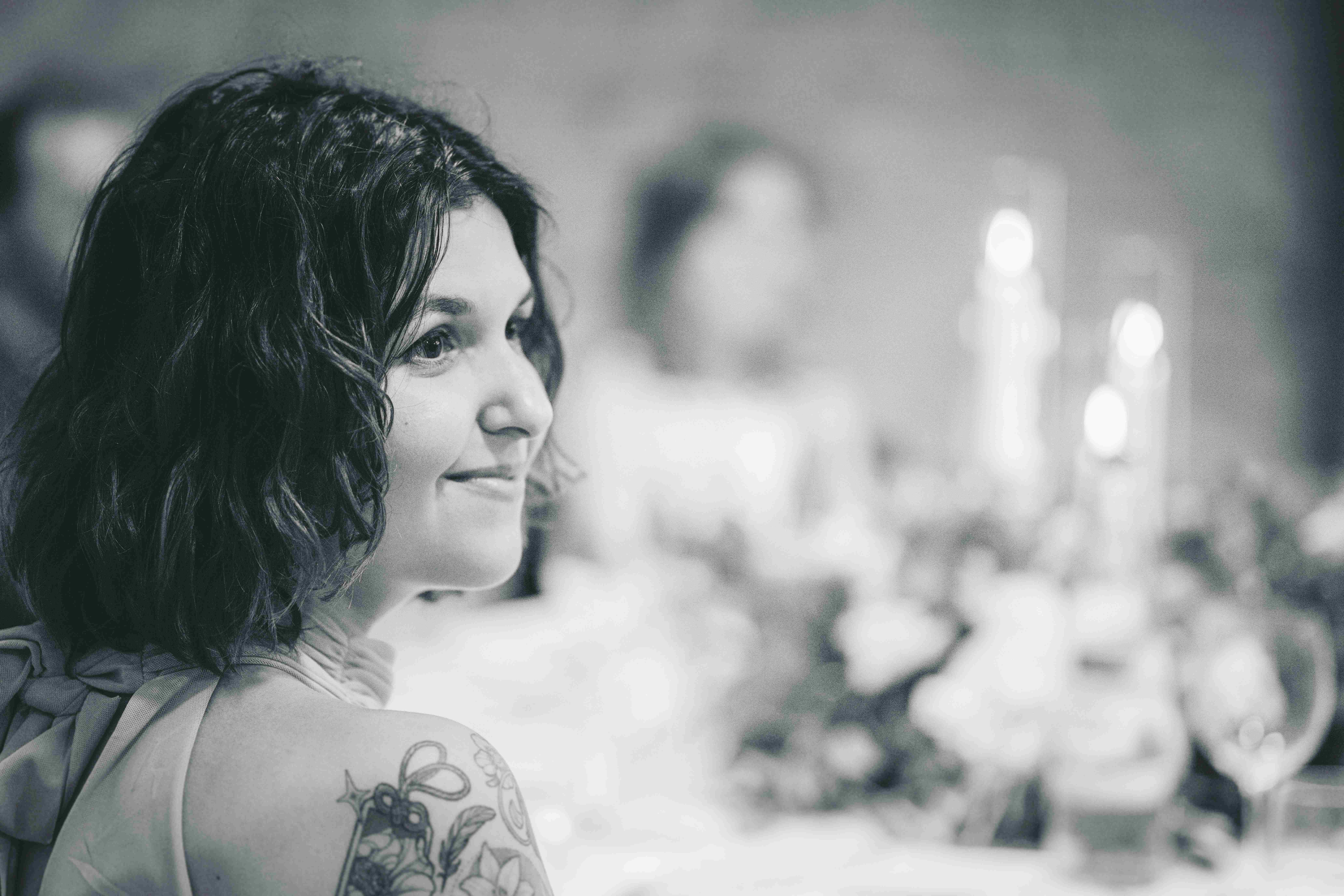 Schwarz-Weiß-Nahaufnahme einer lächelnden Gästin mit dunklen Locken und Tattoo an der festlich dekorierten Hochzeitstafel im Magdalenengut Erfurt.