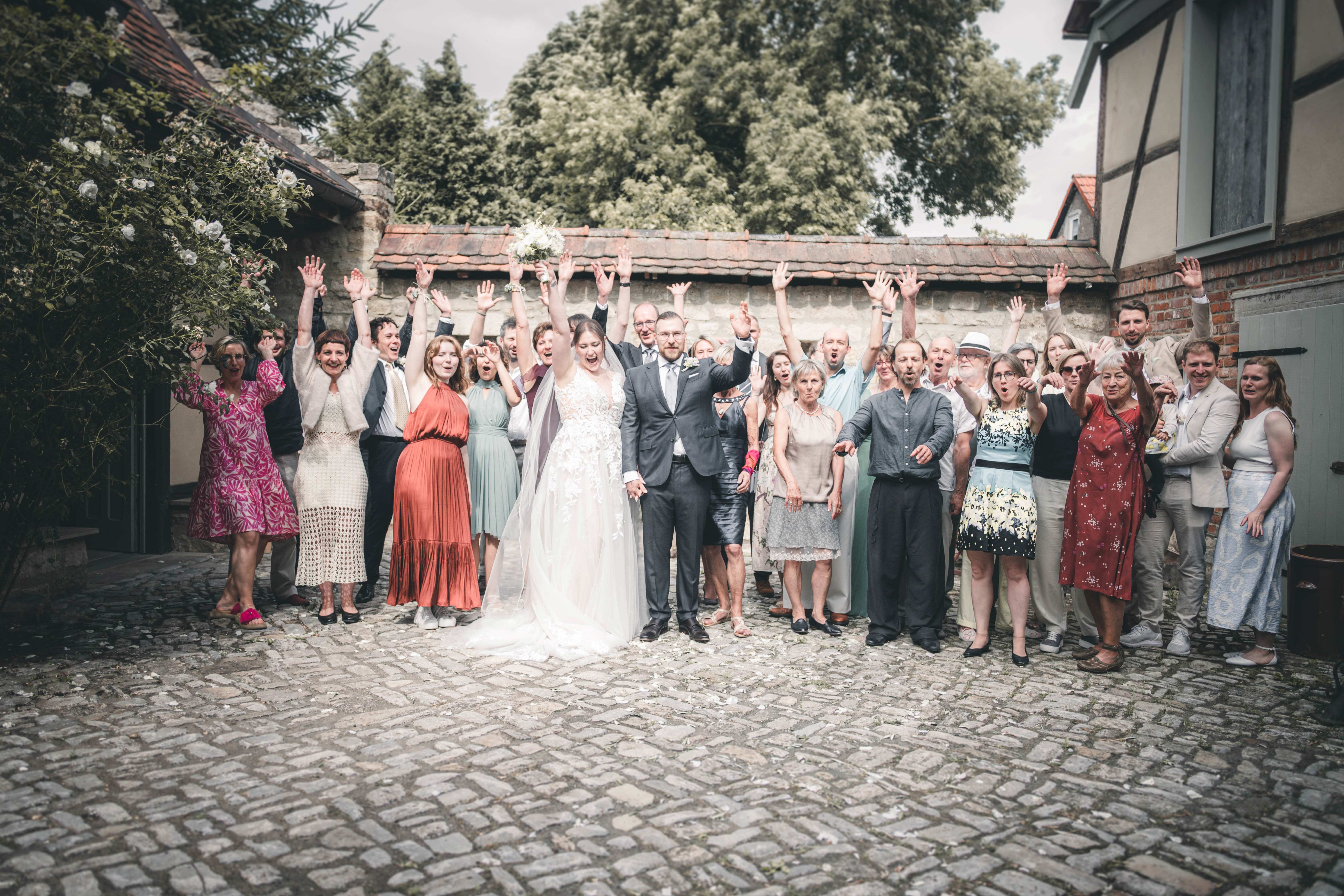 Alle Hochzeitsgäste jubeln mit erhobenen Händen auf dem historischen Kopfsteinpflaster im Innenhof des Magdalenenguts Erfurt.