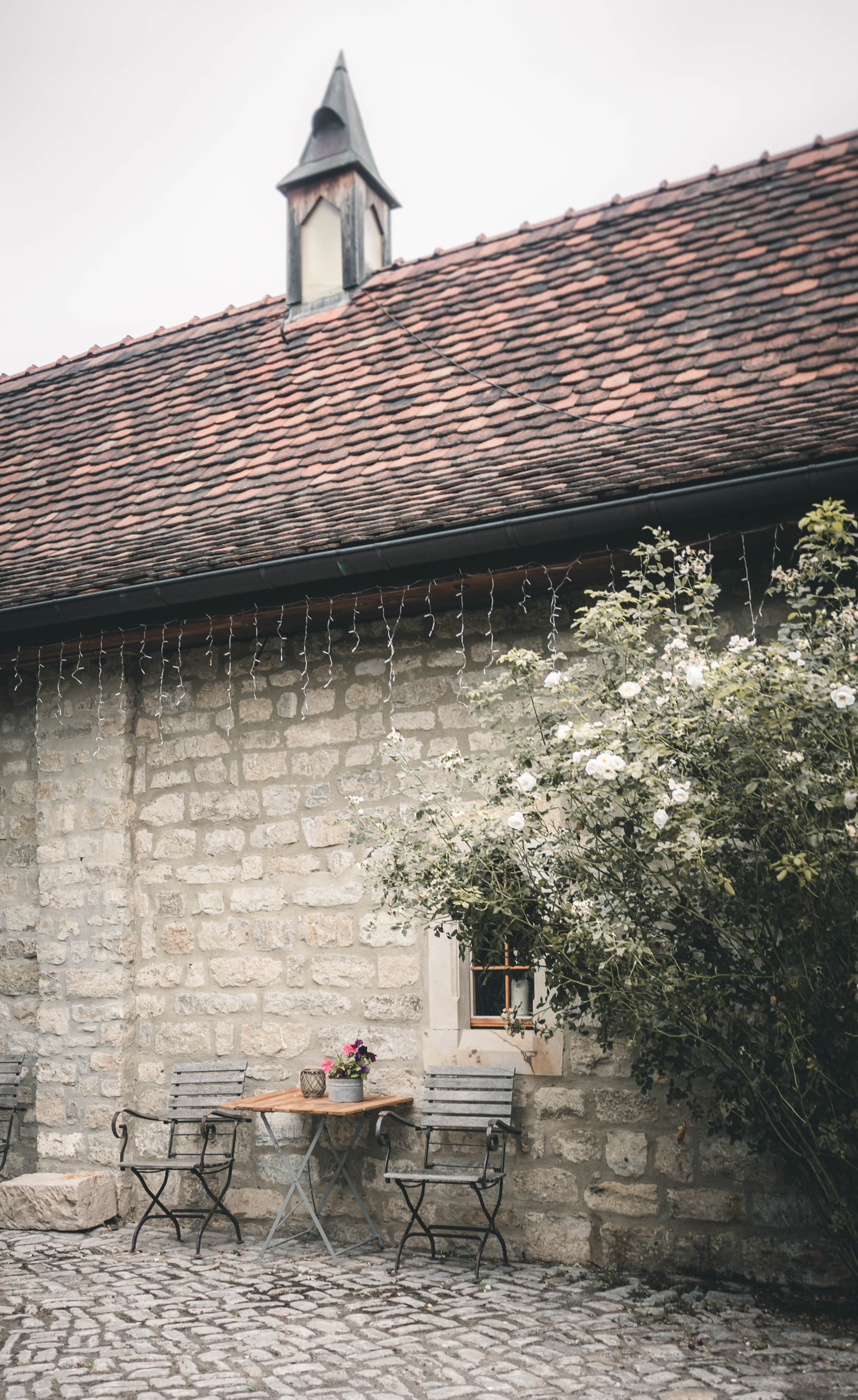 Historische Natursteinmauer der Kapelle im Magdalenengut Erfurt mit Holztisch, schmiedeeisernen Stühlen und blühendem Rosenstrauch.
