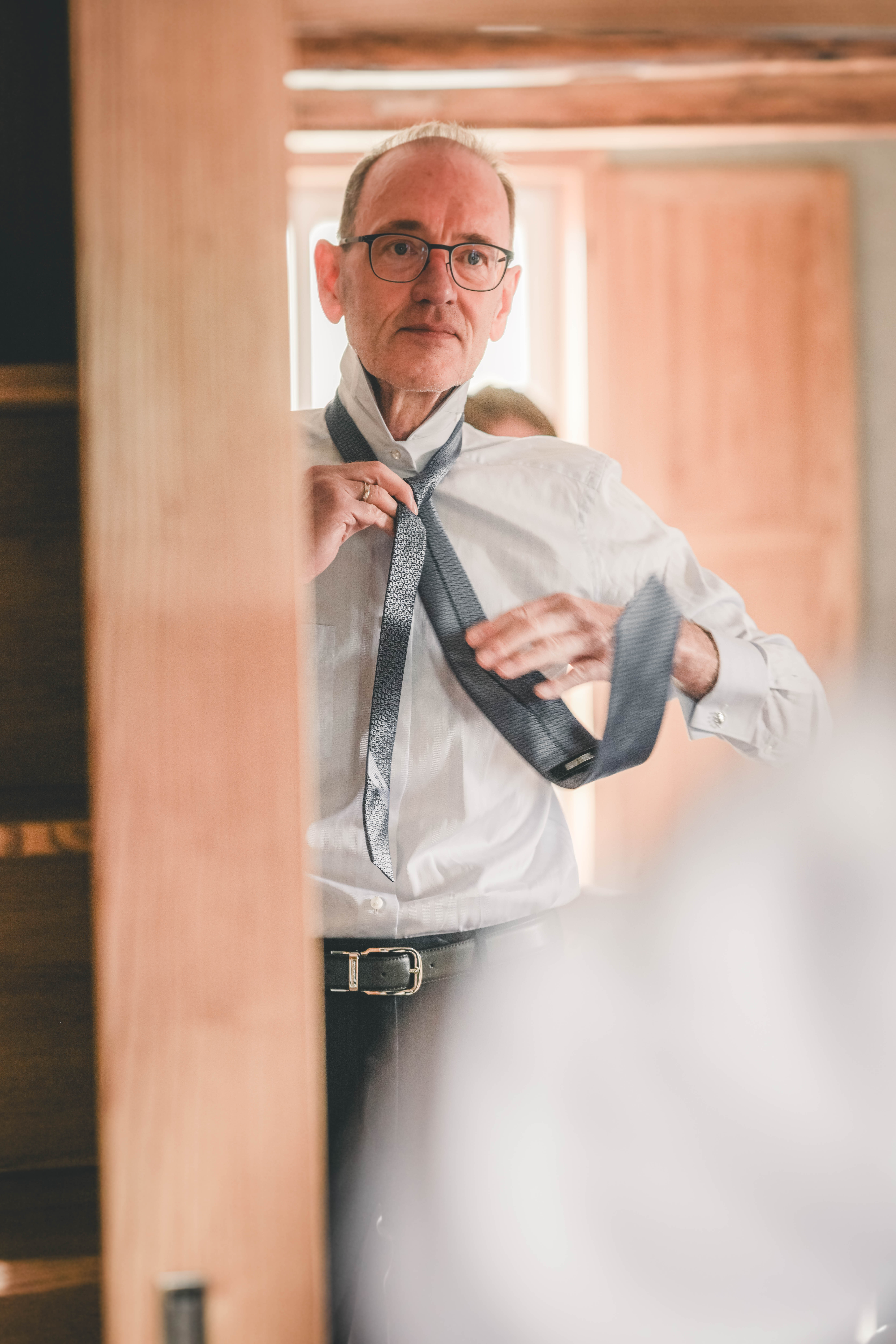 Spiegelaufnahme eines Mannes mit Brille, der sich im Raum des Magdalenenguts Erfurt vor dem Fenster die Krawatte umlegt.