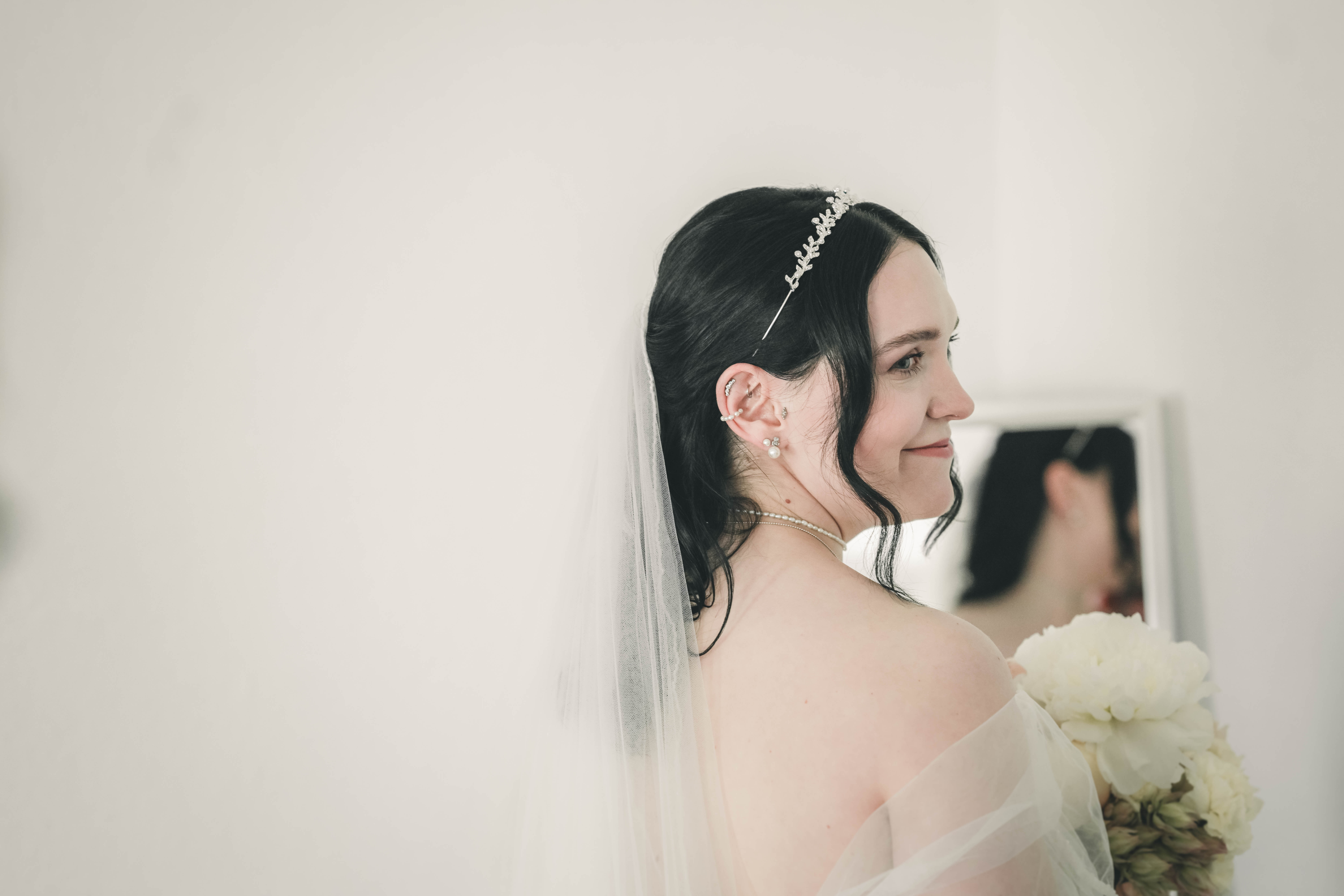 Die Braut steht im fertigen, ausladenden Hochzeitskleid mit zarten Off-Shoulder-Ärmeln aus Tüll im hellen Zimmer im Schloss.