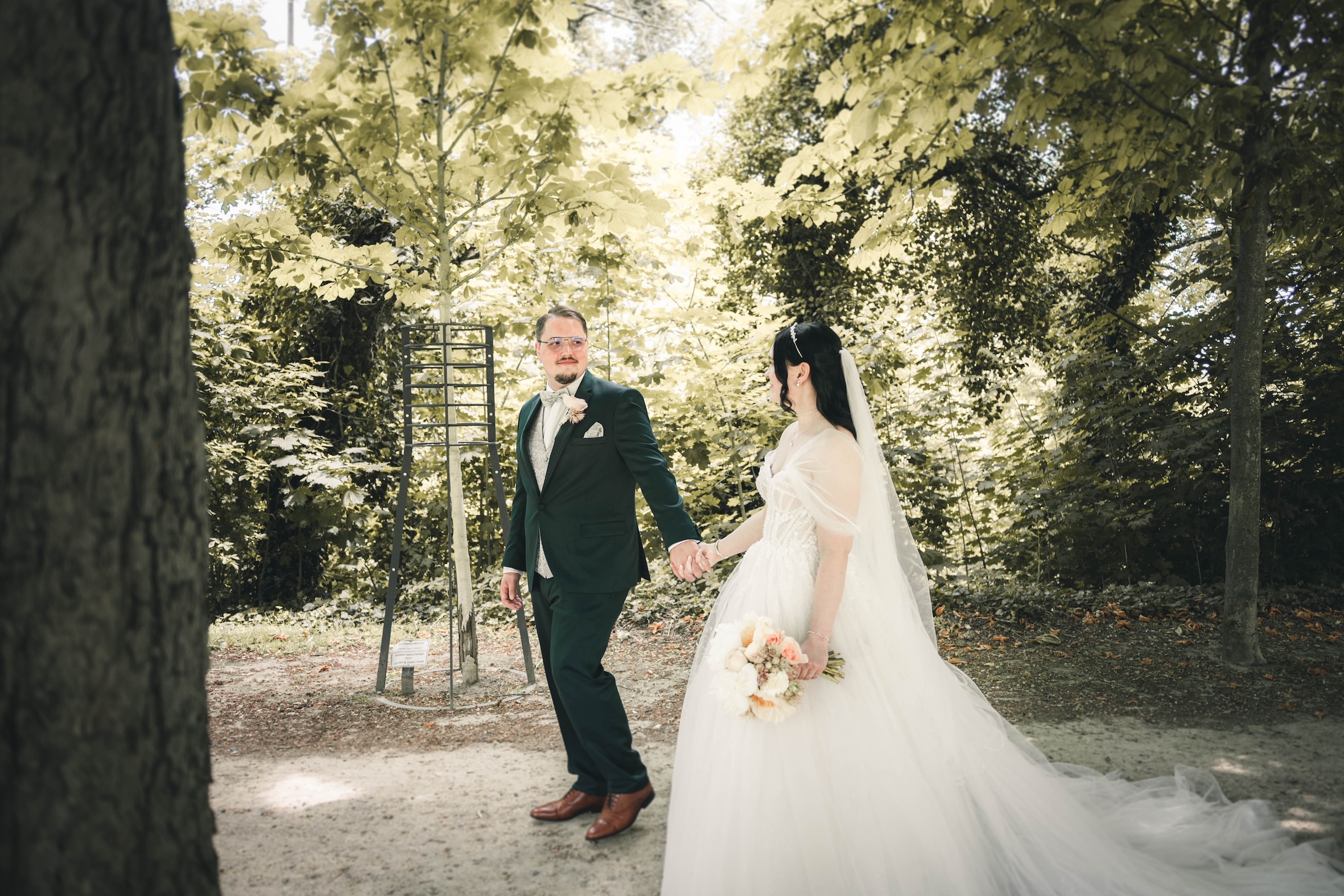 Das Brautpaar läuft Hand in Hand über einen von schattigen Bäumen gesäumten Weg im Schlosspark Frankleben und blickt sich zärtlich an.