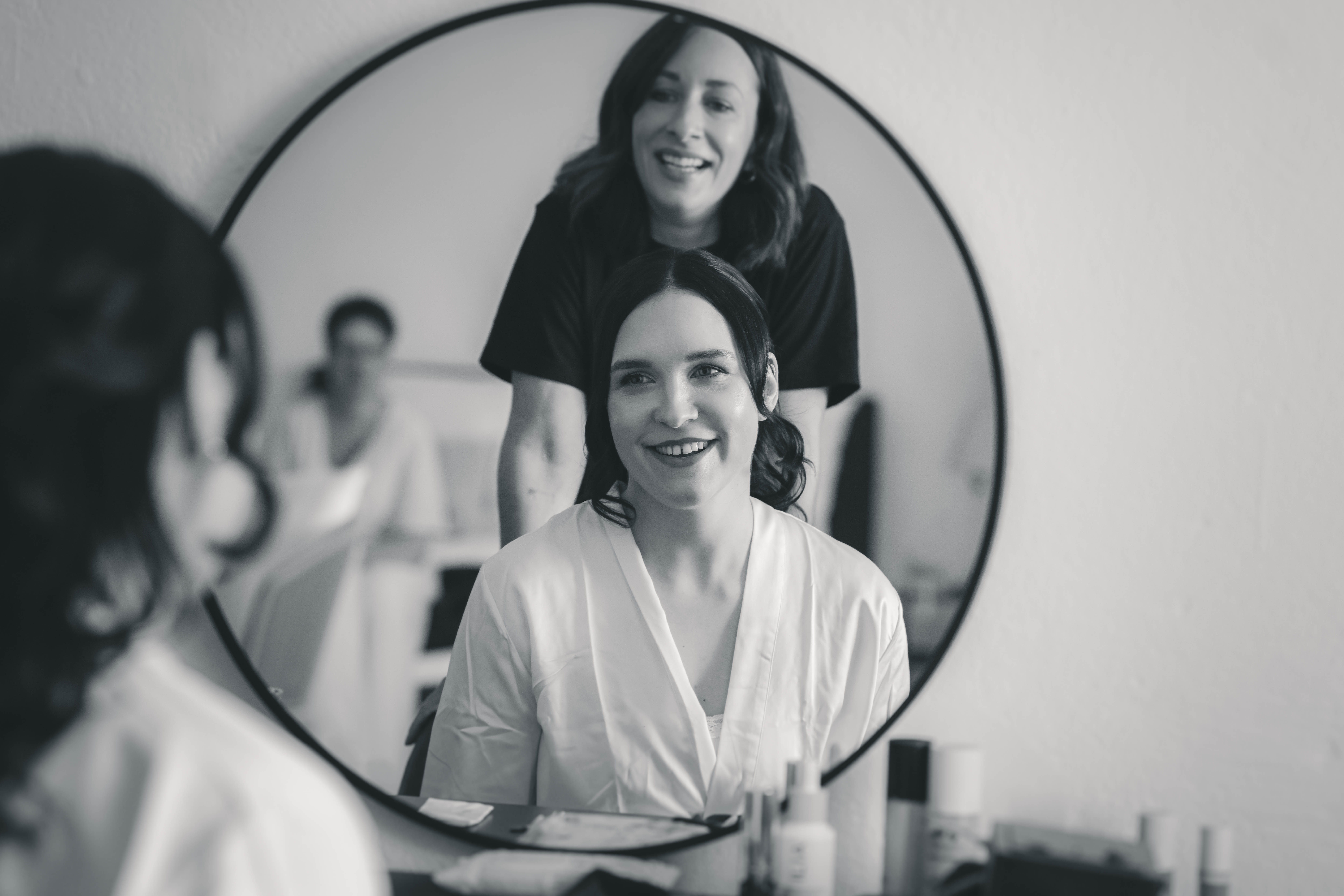 Eine stilvolle Schwarz-Weiß-Spiegelung im runden Kosmetikspiegel zeigt die lächelnde Braut und ihre Make-up-Artistin beim Getting Ready.