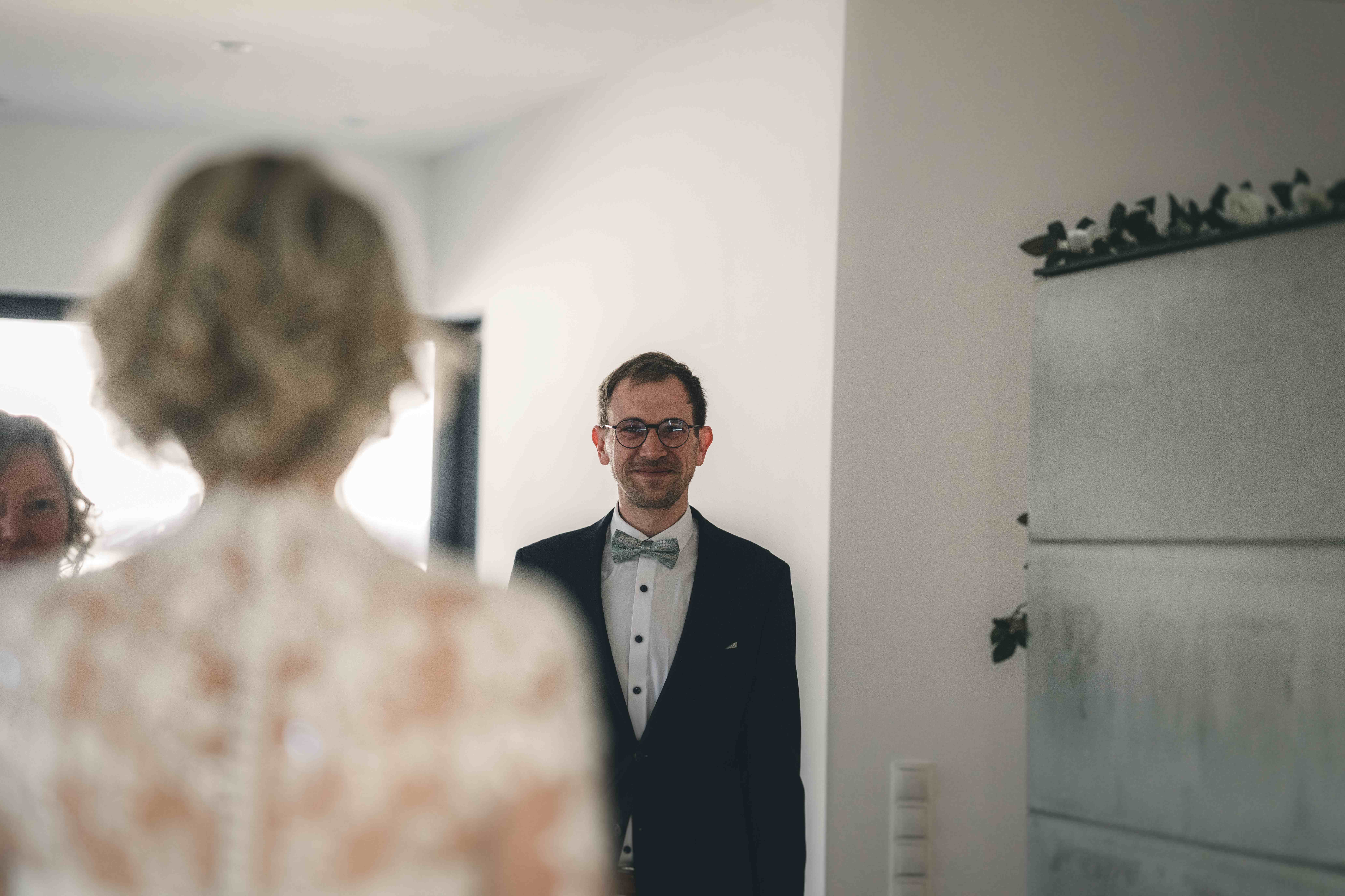 Der Bruder im schwarzen Anzug blickt seine Schwester beim intimen First Look im Haus in Fraureuth mit einem emotionalen, glücklichen Lächeln an.