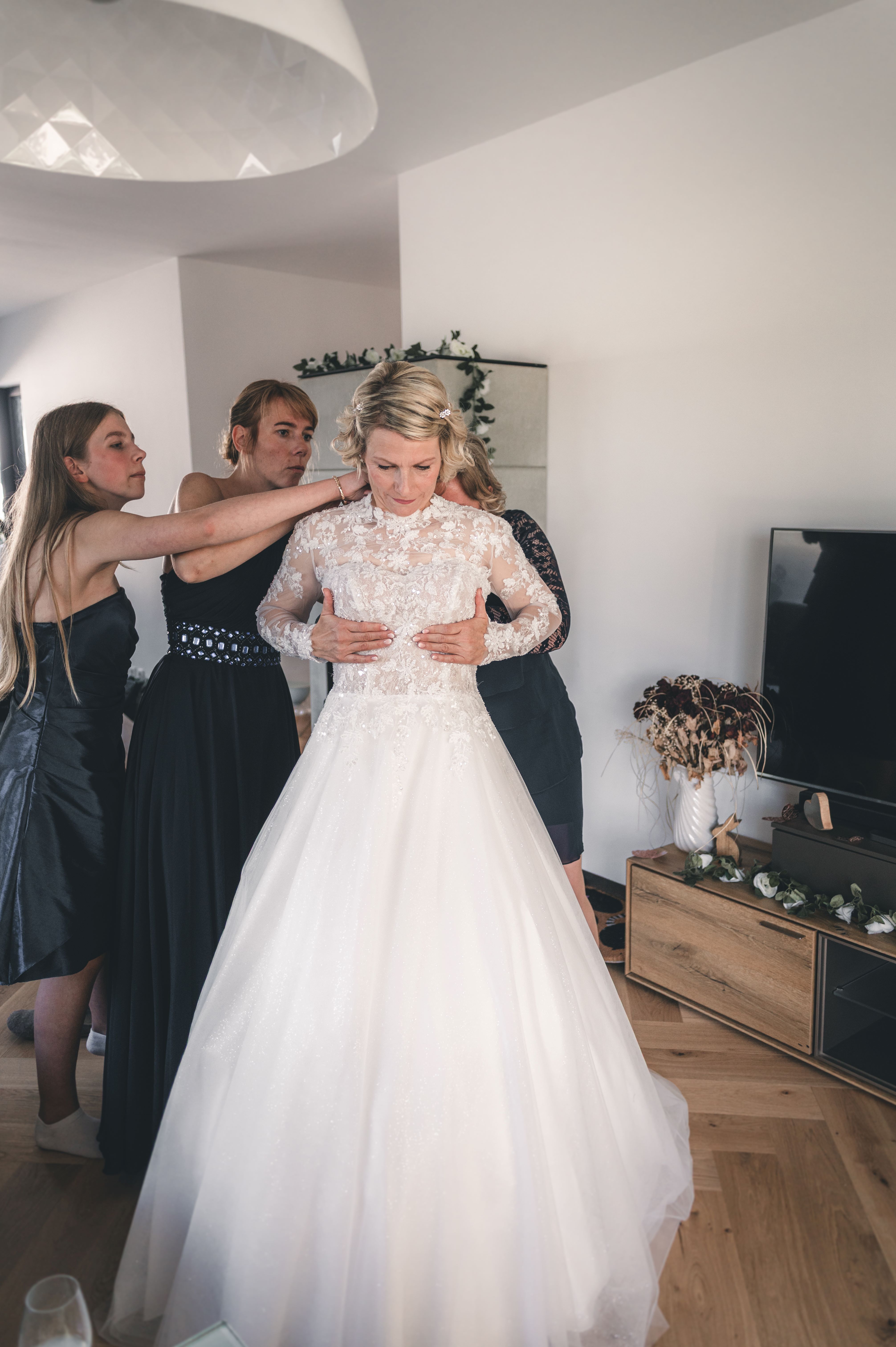 Das Brautkleid sitzt perfekt: Drei Freundinnen legen letzte Hand an das hochgeschlossene Spitzenkleid der Braut in Fraureuth, Erzgebirge.