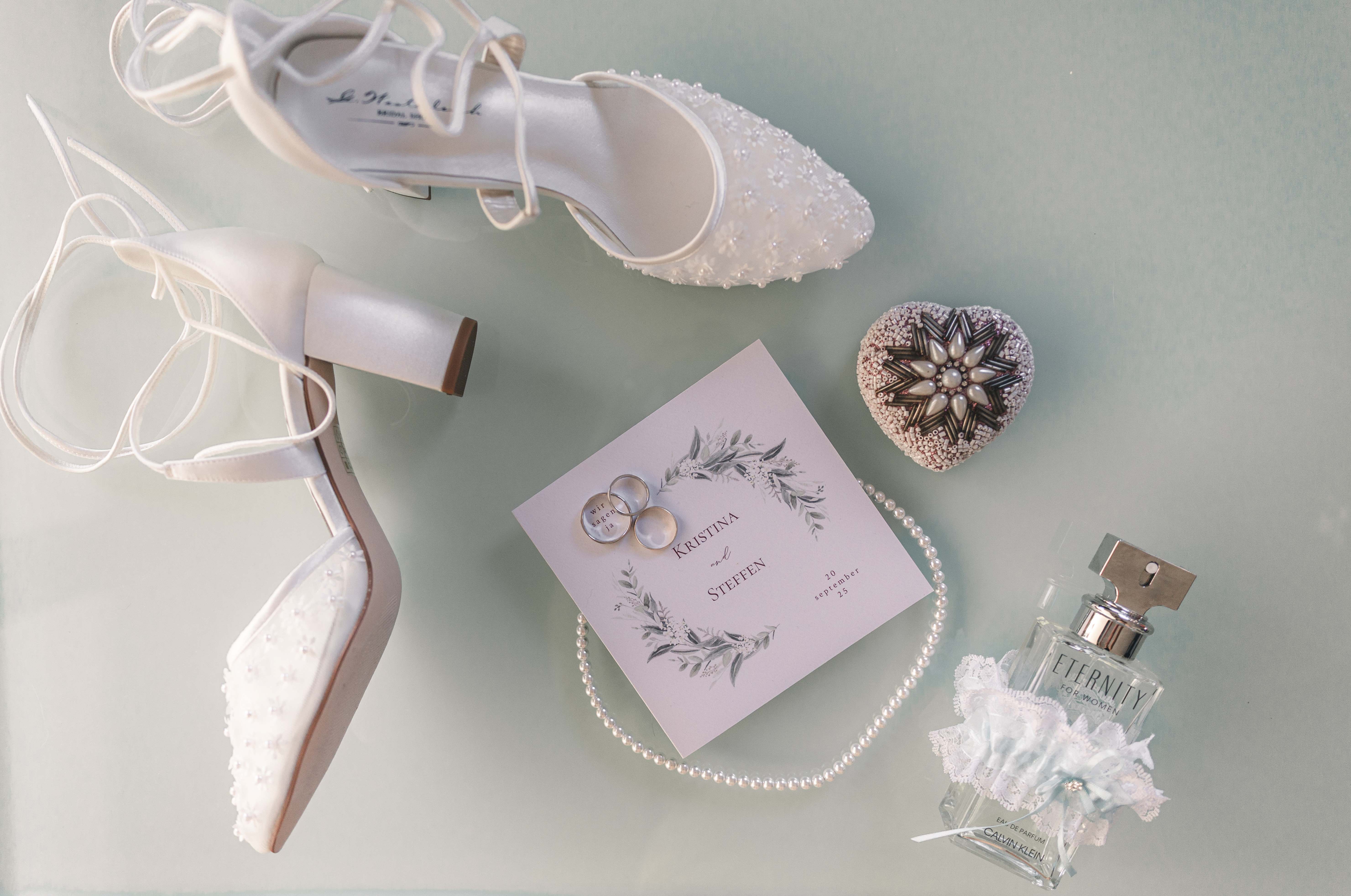 Eine edle Flatlay-Aufnahme von Brautschuhen mit Spitze, den Eheringen auf der Einladungskarte, einer Perlenkette und Parfum vor der Trauung in Fraureuth.