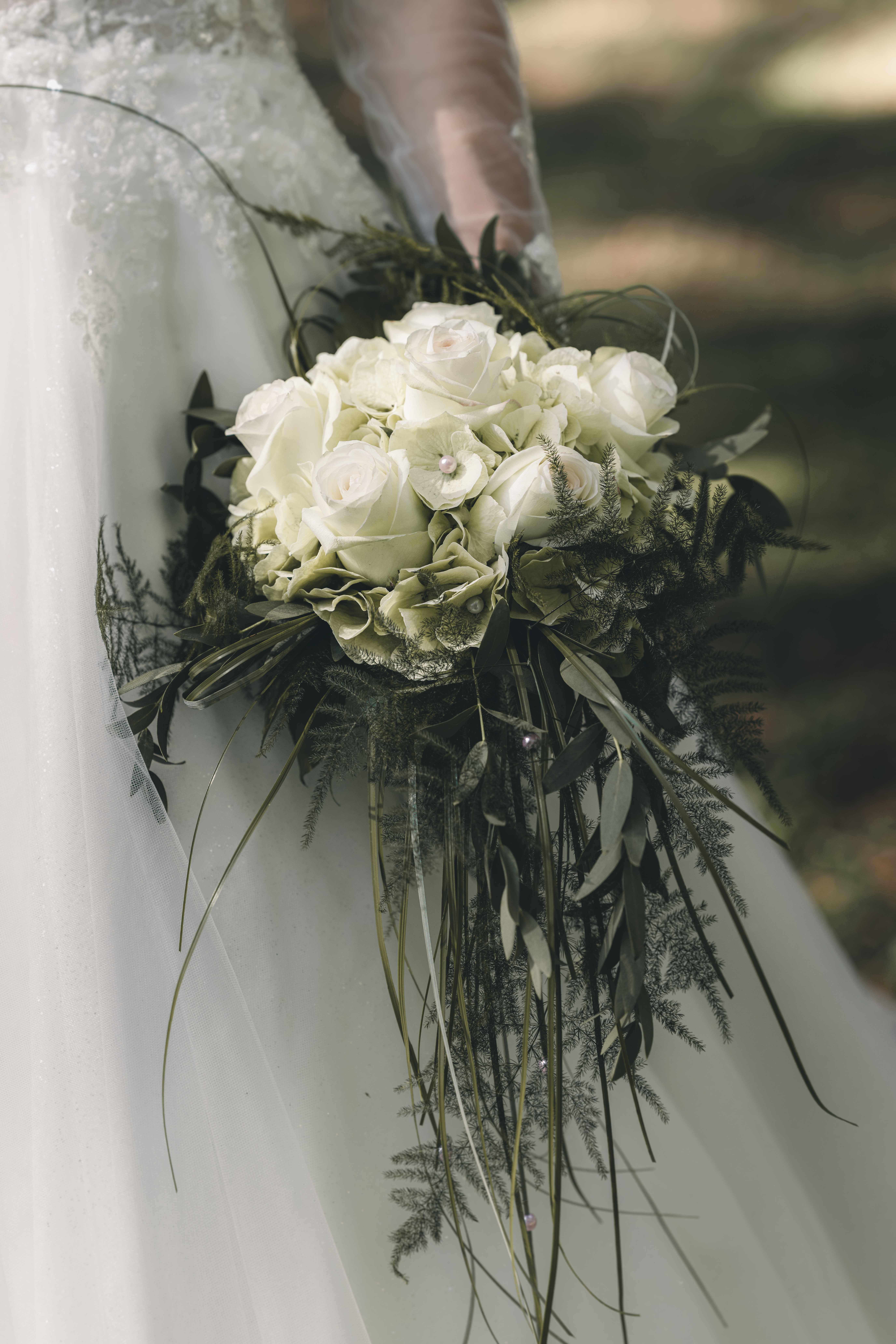 Detailaufnahme des eleganten, fließenden Brautstraußes aus weißen Rosen, feinen Perlen und grünem Farn vor dem weißen Tüll des Kleides.