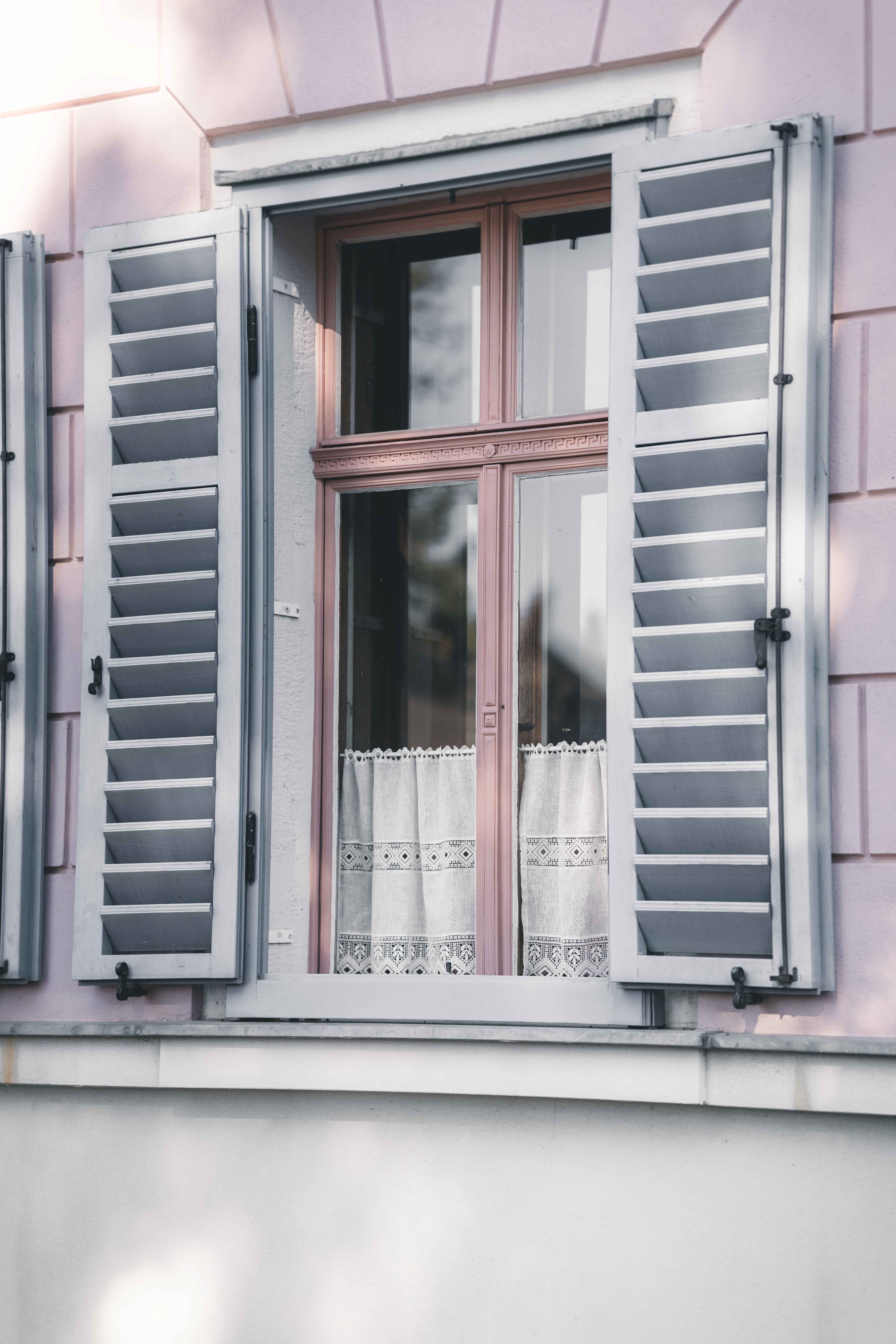 Ein geschlossenes Fenster mit weißen Spitzenvorhängen und geöffneten, grauen Holzfensterläden an der rosa Fassade des Standesamts Fraureuth.