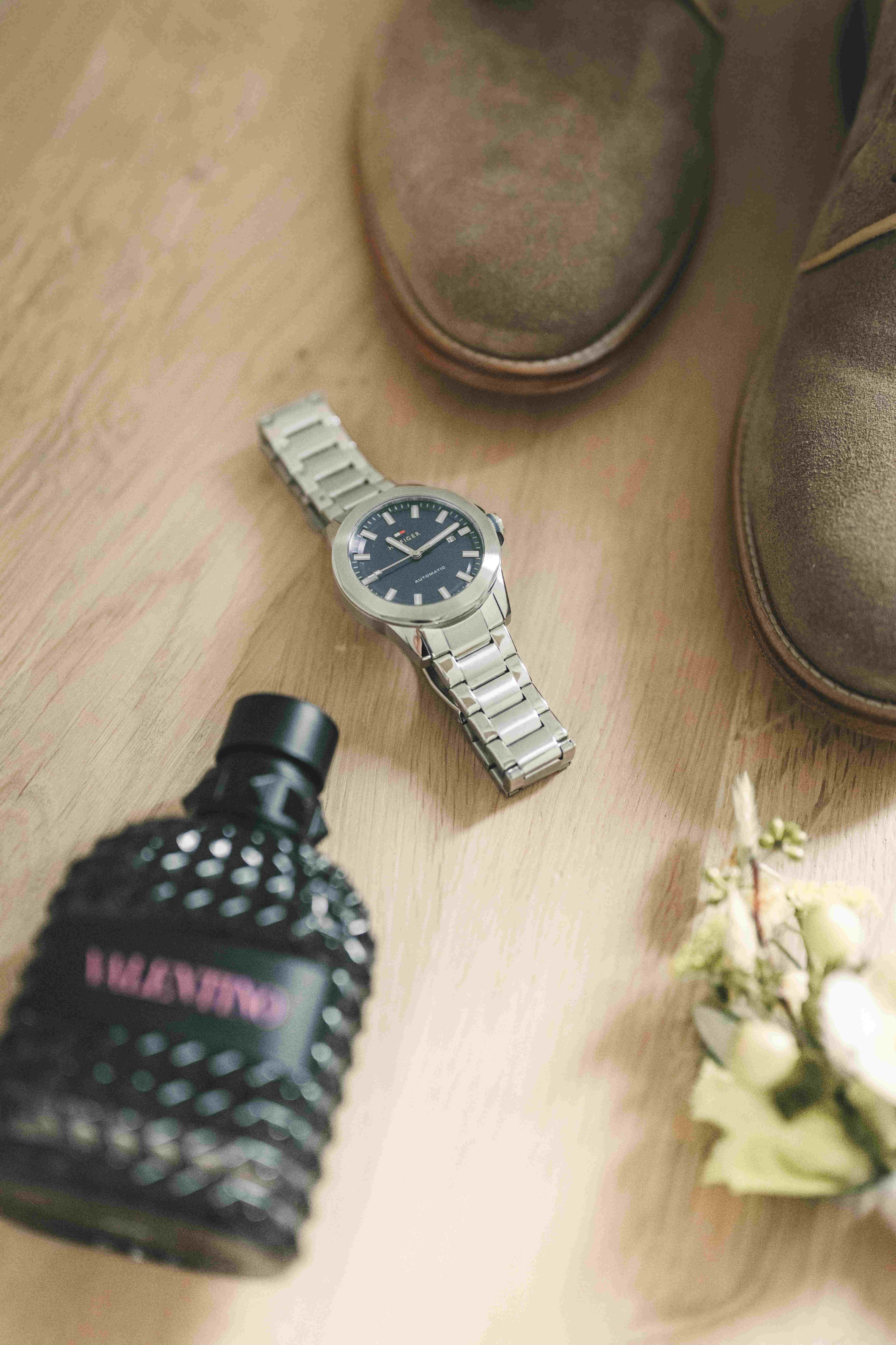 Flachliegende Hochzeitsdetails des Bräutigams auf Holzboden: Eine elegante silberne Herrenuhr, Valentino-Parfüm, Wildlederschuhe und eine Ansteckblume.