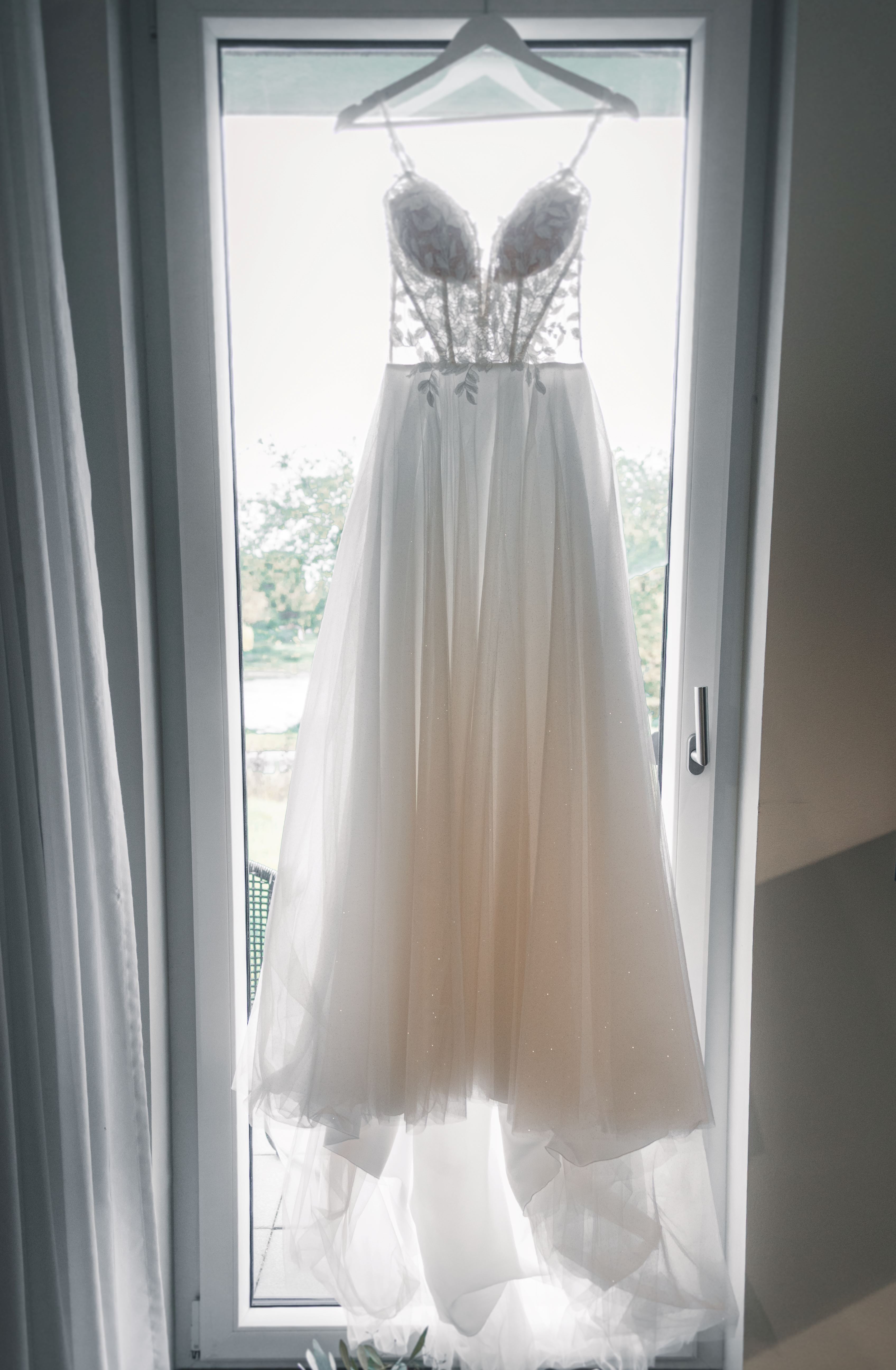 Ein wunderschönes, filigranes Brautkleid mit Spitzen-Korsage und fließendem Tüll-Rock hängt an einer großen Fenstertür im hellen Vorbereitungszimmer.